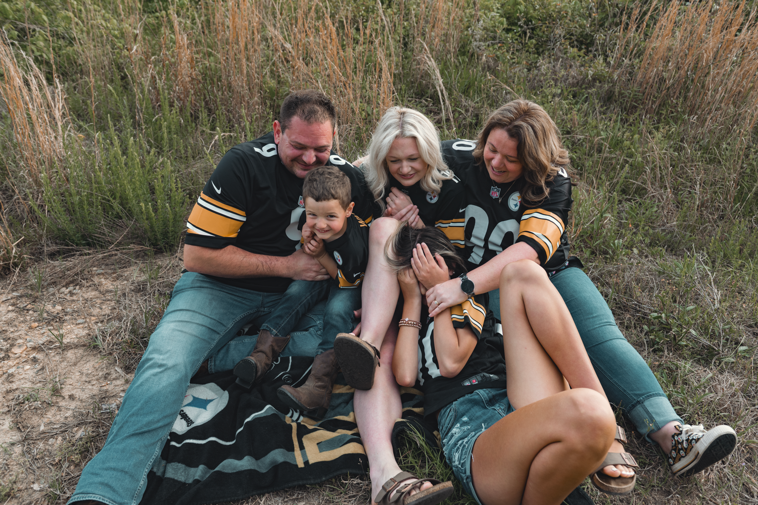 Three people in football jerseys laugh together while relaxing on a blanket in a field with tall grass at sunset.