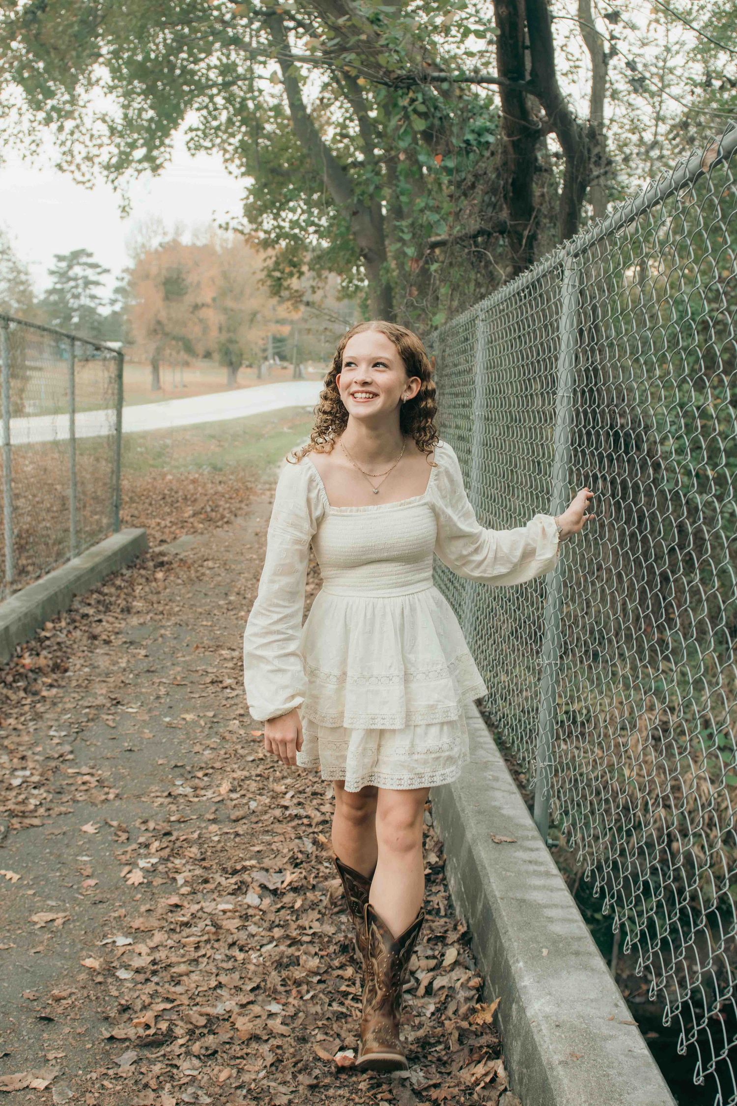 Someone in a white prairie dress and brown cowboy boots walks along a tree-lined path with a chain link fence during fall in Smithfield, NC
