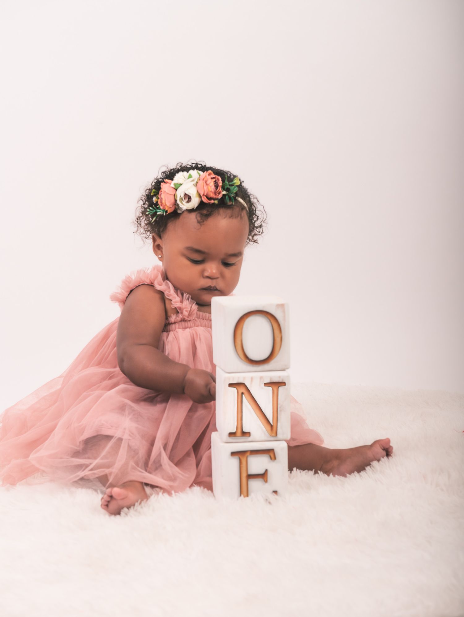 Baby in a pink tulle dress and floral crown sitting beside wooden blocks spelling 'ONE' on white backdrop.