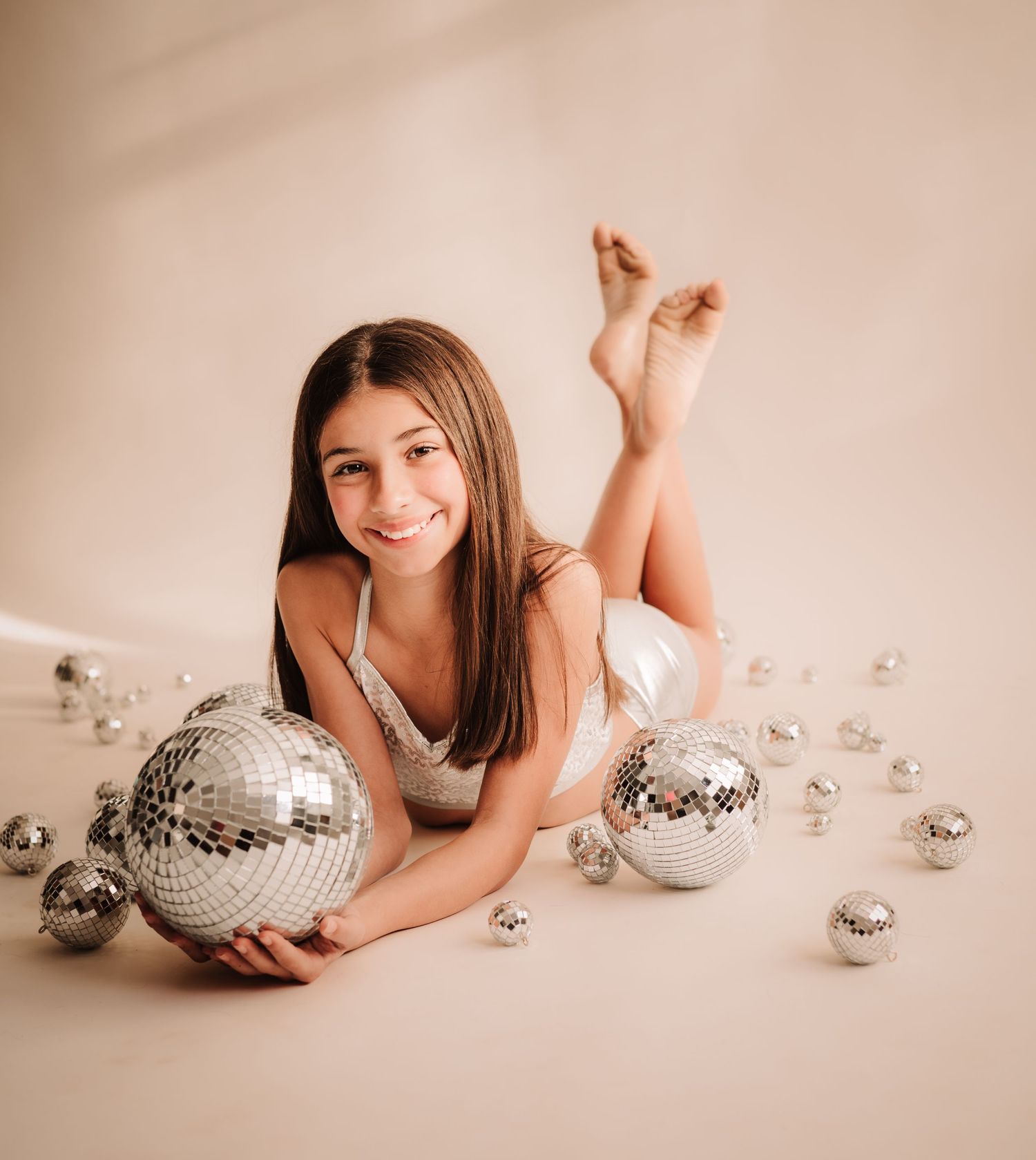 A person poses with silver disco balls on a beige background during a glamorous photoshoot.