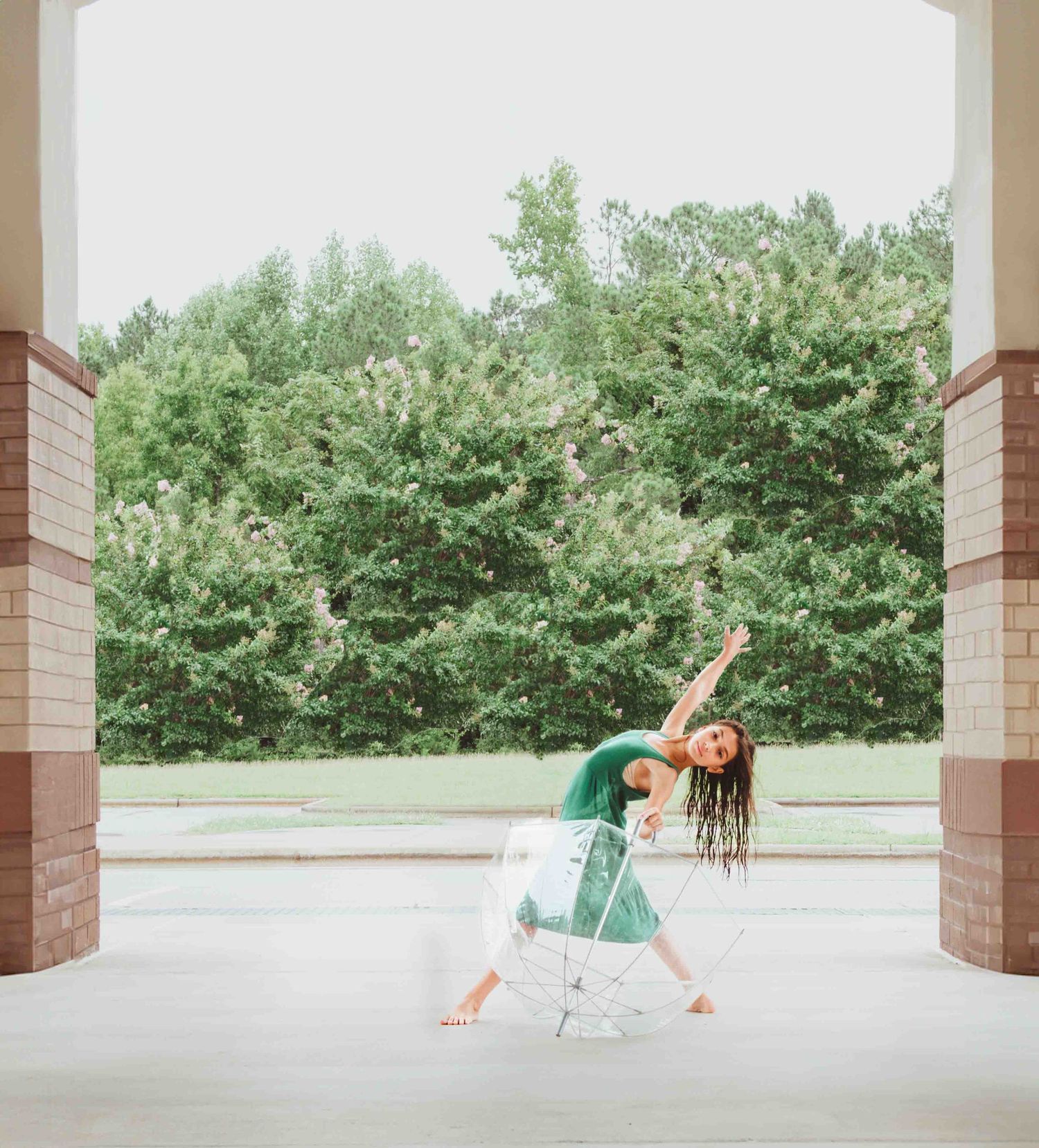 A dancer in a green outfit performs a graceful pose under a covered walkway with trees in the background.
