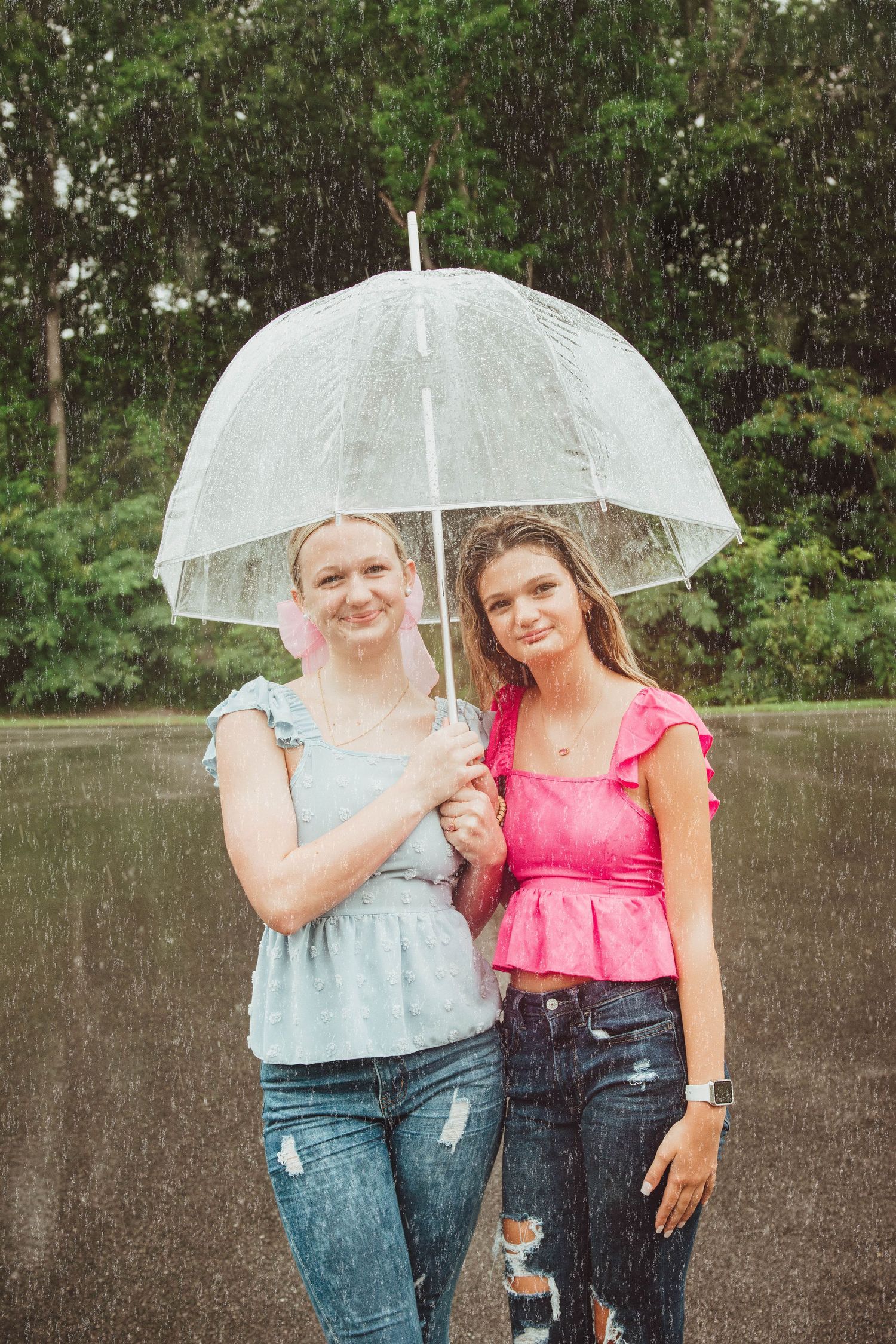 Two people stand together under a clear bubble umbrella in the rain while wearing casual summer outfits.