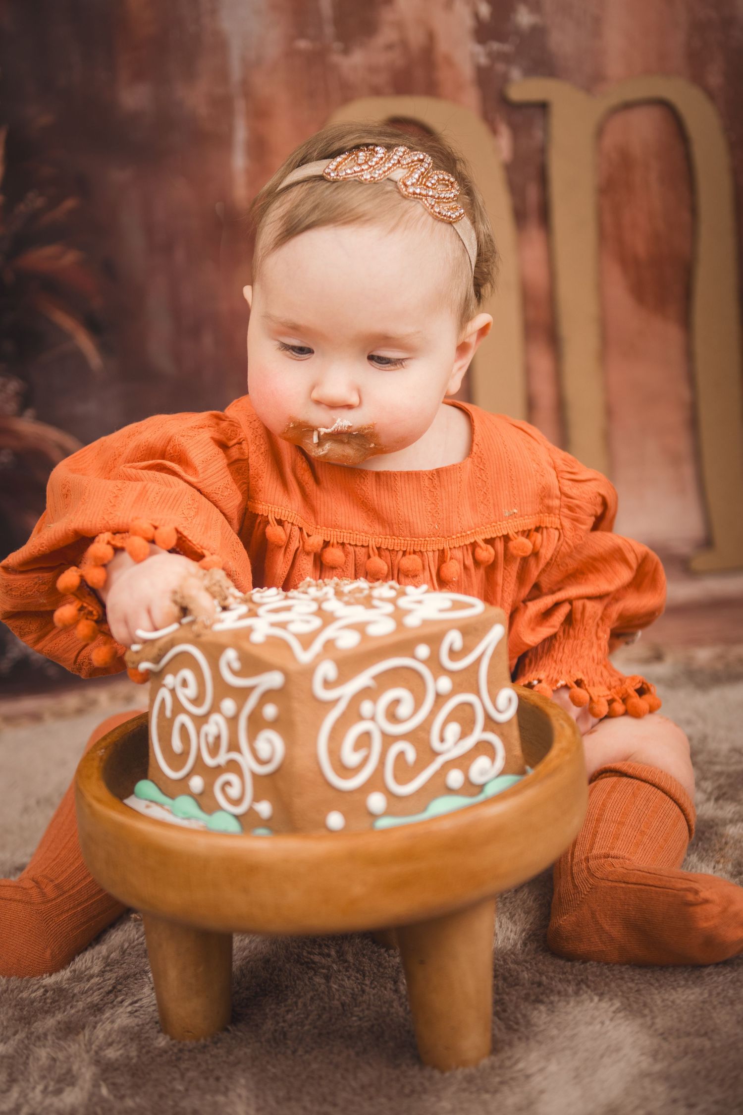 Baby in orange dress sits with decorated brown and white cake on small wooden stool during autumn photo session.