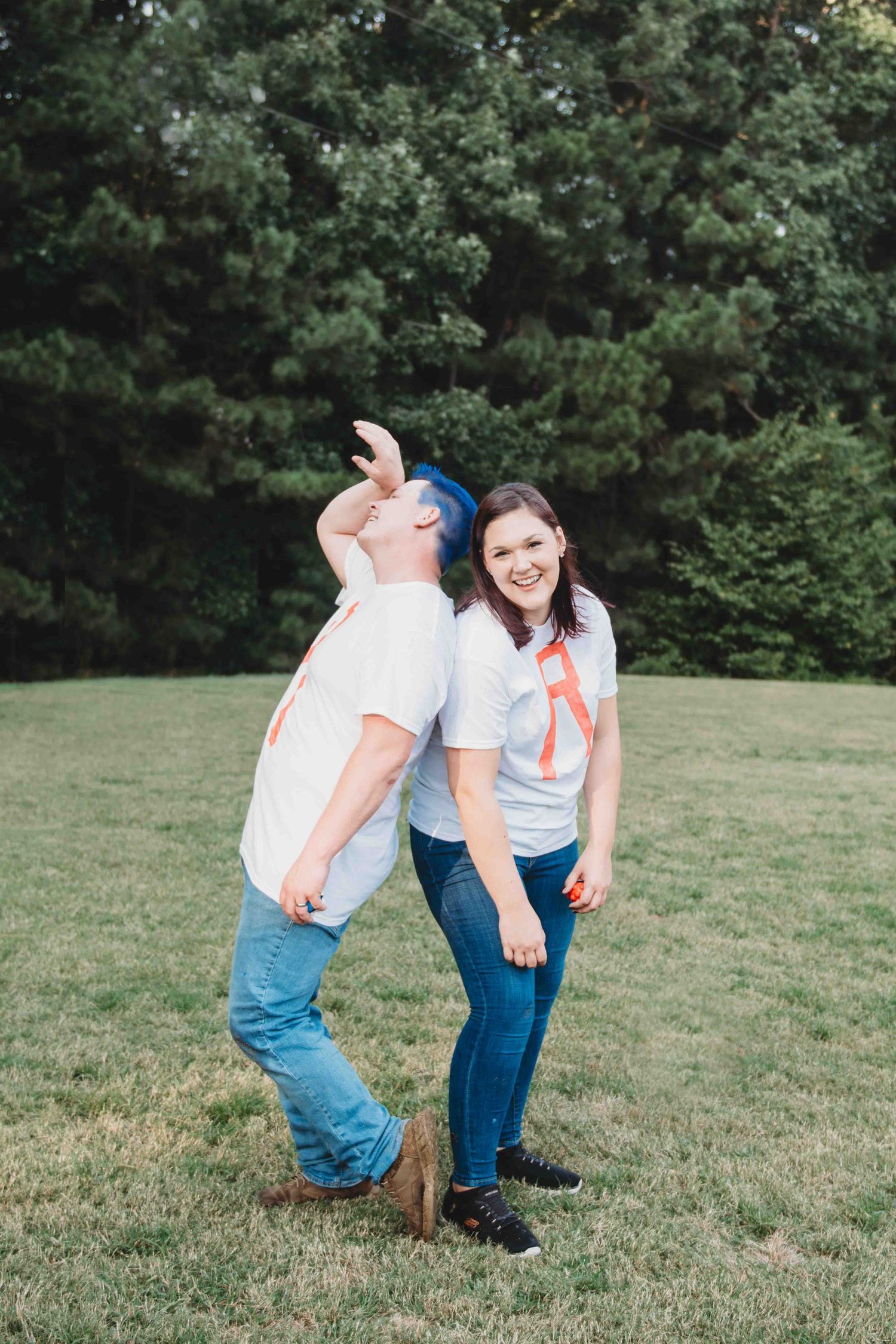 A couple poses playfully together on a grassy hill with trees in the background.