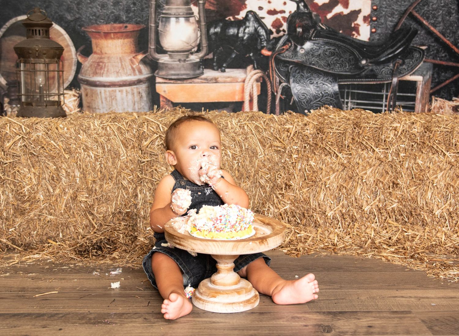 A baby in overalls sits on wooden floor against hay bales with a rustic cake stand and farmhouse decor in background.