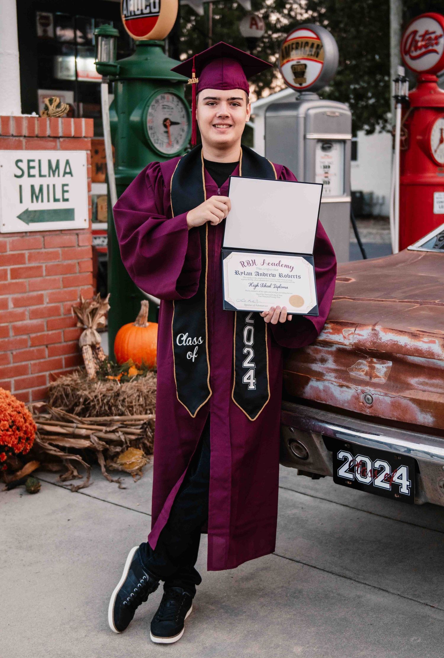 Graduate in maroon cap and gown holds diploma while posing near vintage gas pumps and fall decorations.