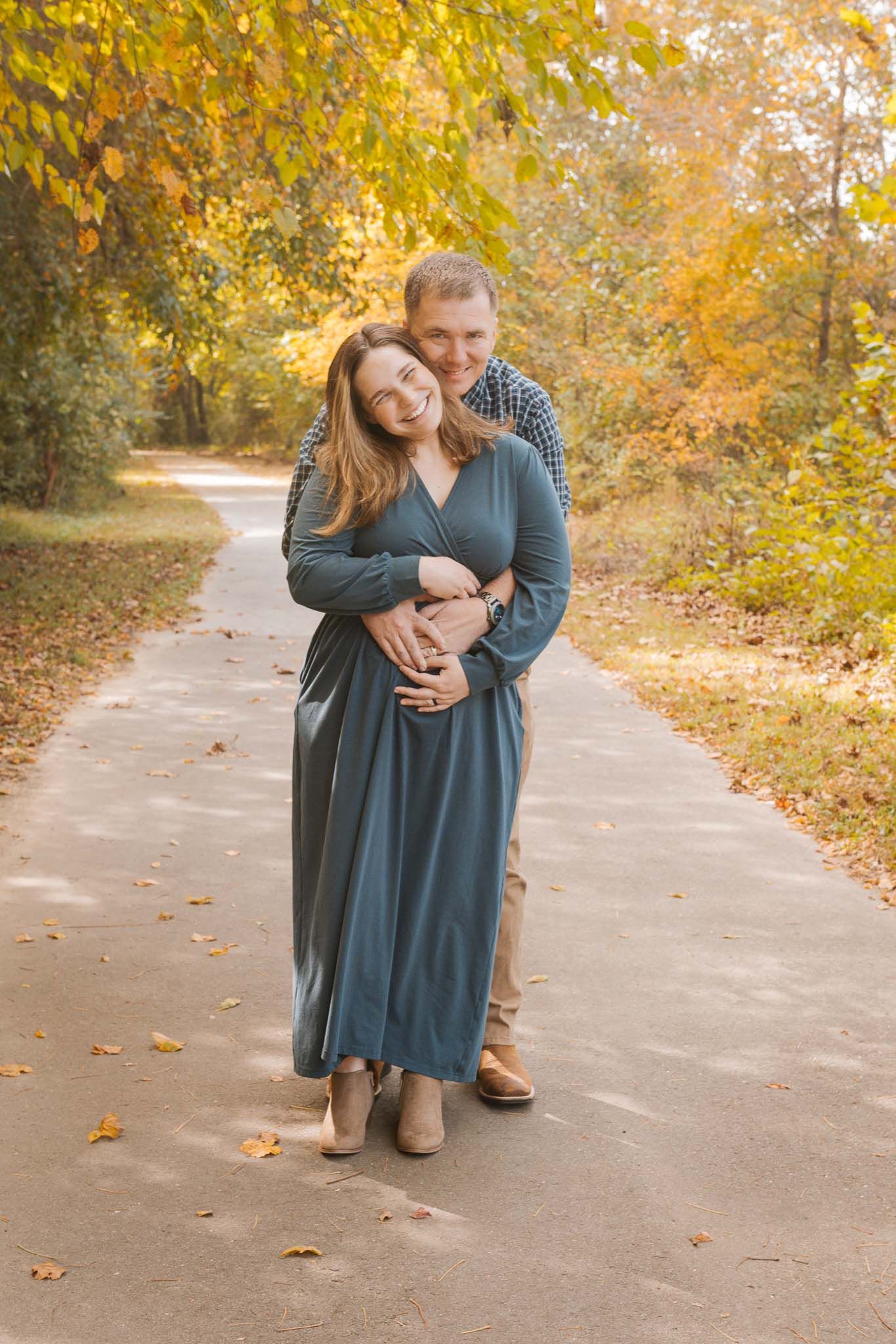 A couple embraces on a path surrounded by golden autumn foliage as sunlight filters through the trees.