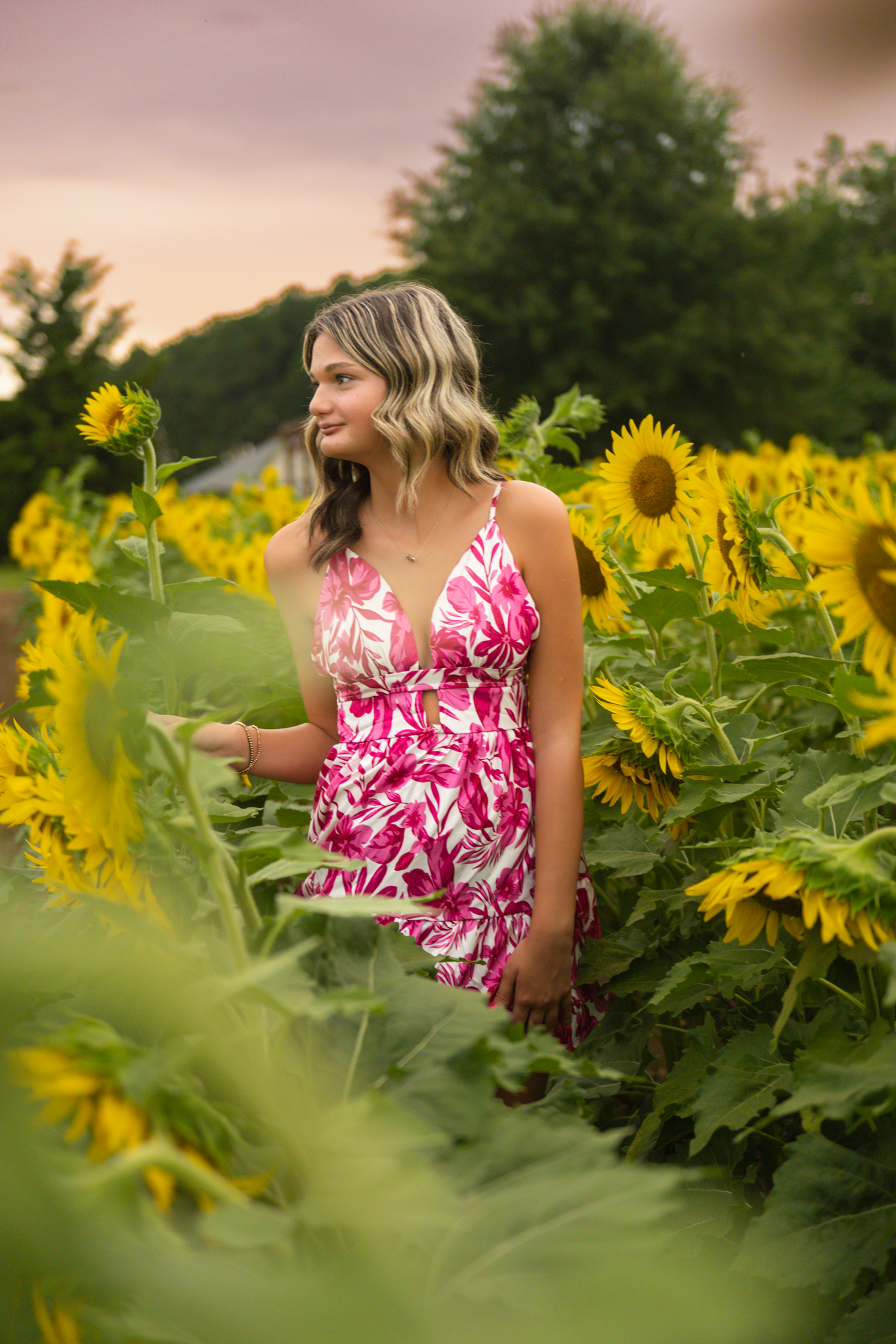 A woman in a pink and white floral dress stands thoughtfully among tall blooming sunflowers during golden hour.