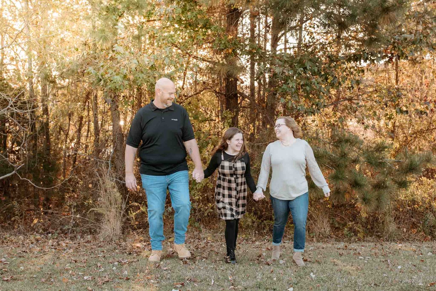 Three people walk together holding hands on a path with autumn foliage and sunlit trees in the background.