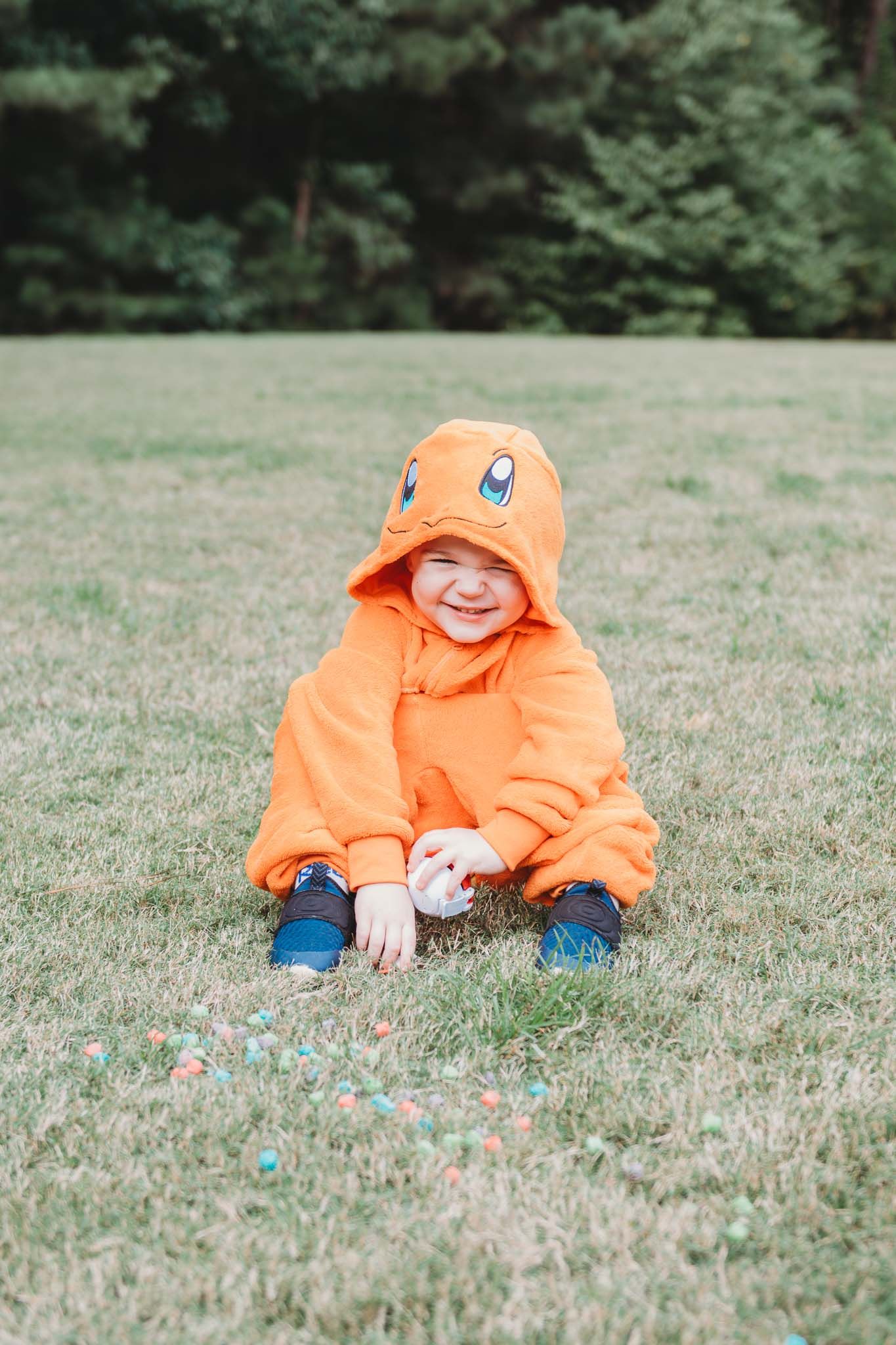 Small figure in bright orange hooded jacket sits joyfully on grass in a park setting.