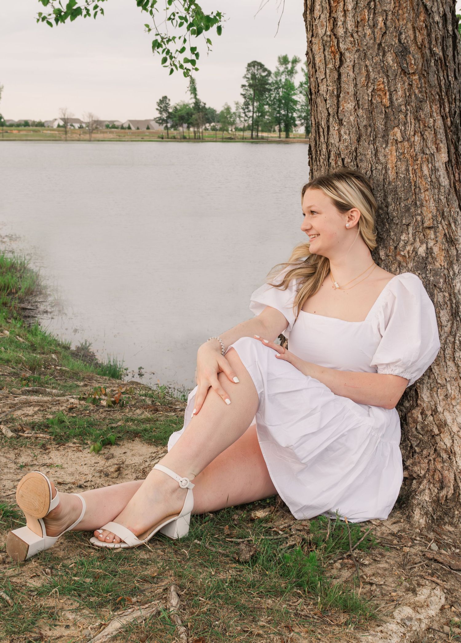 Woman in white dress relaxing against tree trunk by serene lake wearing heeled sandals on summer afternoon.