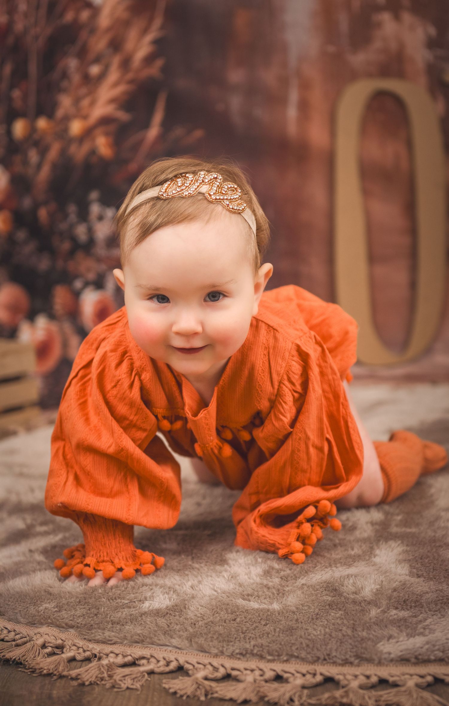 A small child in a bright orange ruffled outfit crawls on a gray carpet in a studio setting.