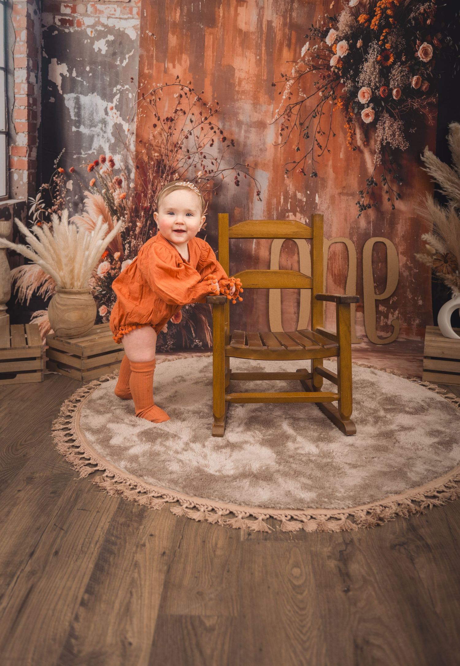 A small child in orange attire stands beside a wooden chair on a round rug in an autumnal-themed studio setting during her 1-year session