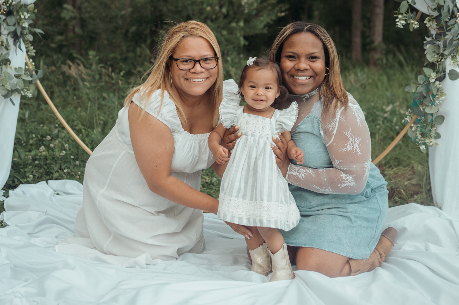 Family seated together on white blanket outdoors during summer photoshoot in garden setting.