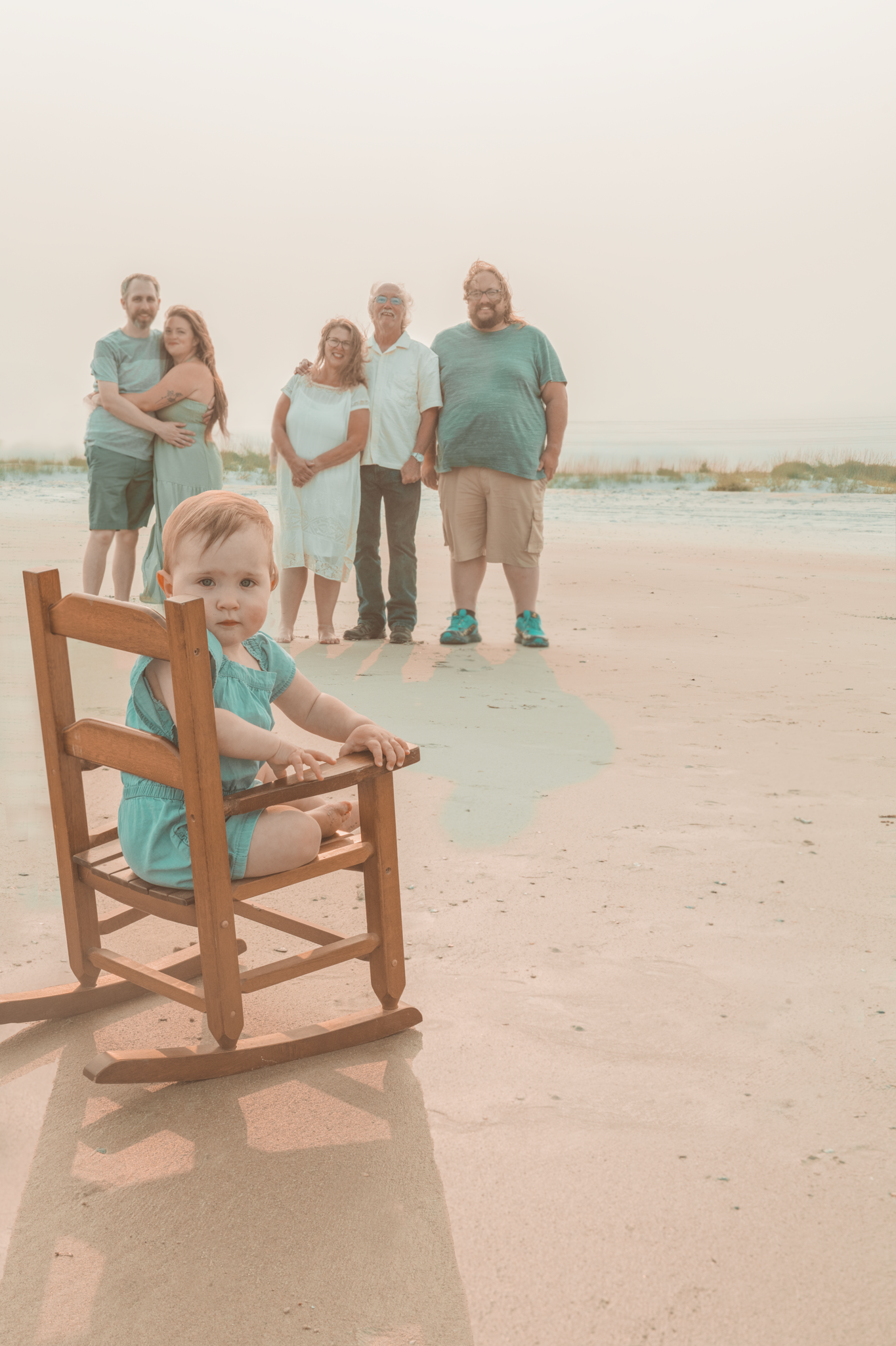 A wooden rocking chair sits on the beach with a family group gathered behind it.