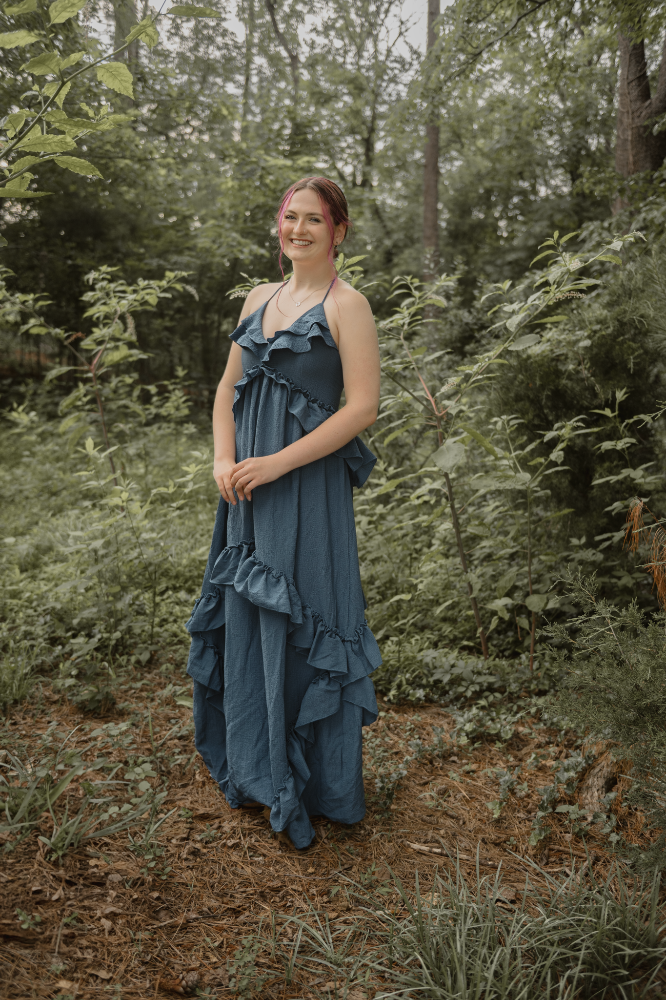 Haylee wearing a flowing blue ruffled maxi dress poses on a forest path surrounded by lush green foliage for her senior session
