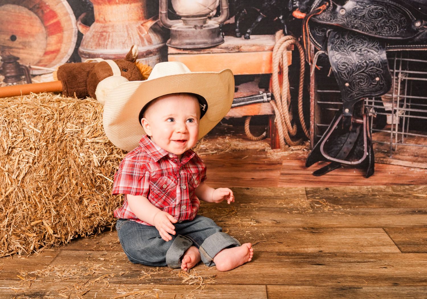 A small cowboy in red plaid shirt and hat sits on wooden floor next to hay bales in rustic country setting.