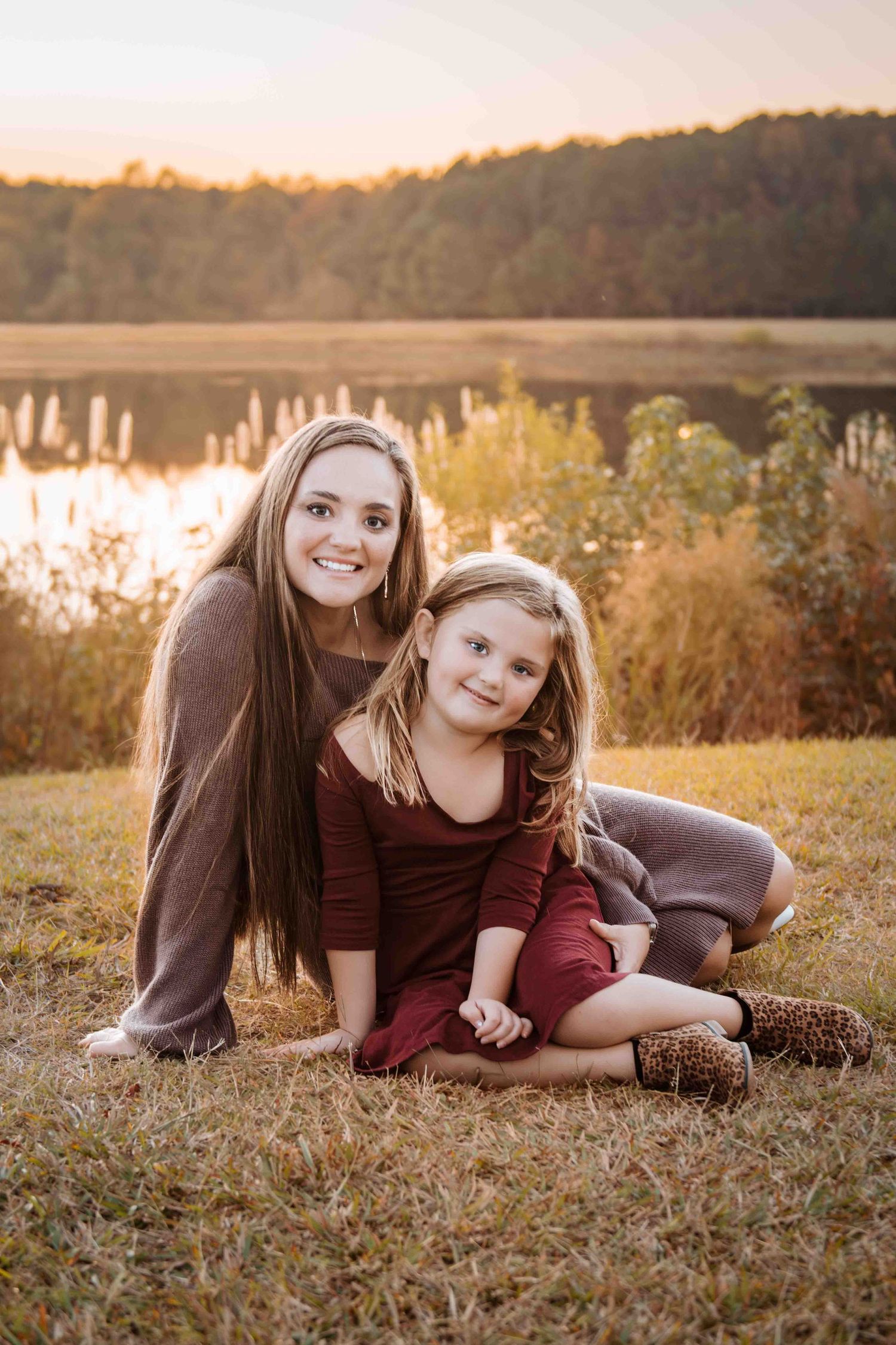 Two young females in burgundy outfits pose together sitting on grass by a lake at sunset in a natural setting.