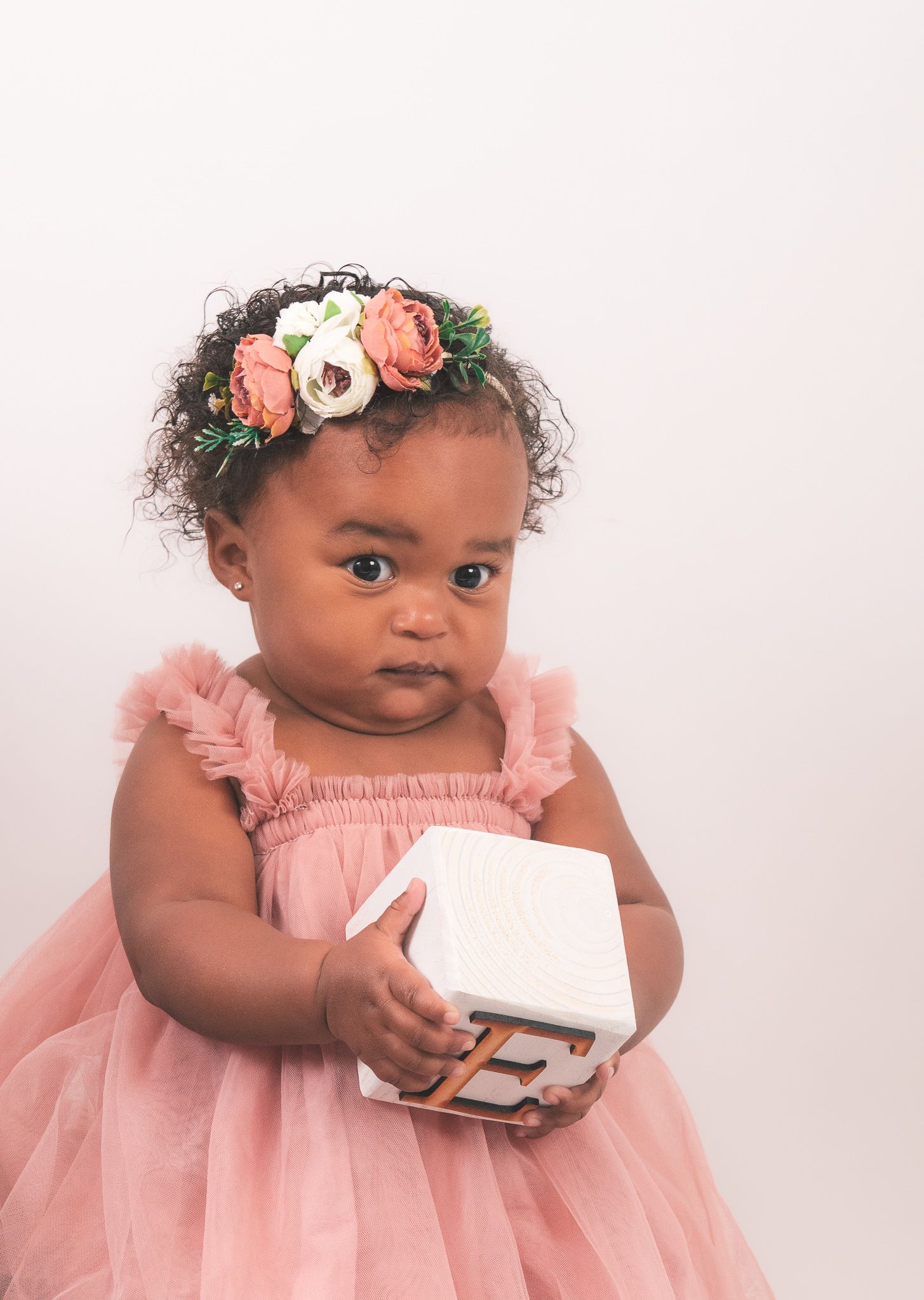 A baby wearing a pink tulle dress and floral crown holds a white gift box against a light background.