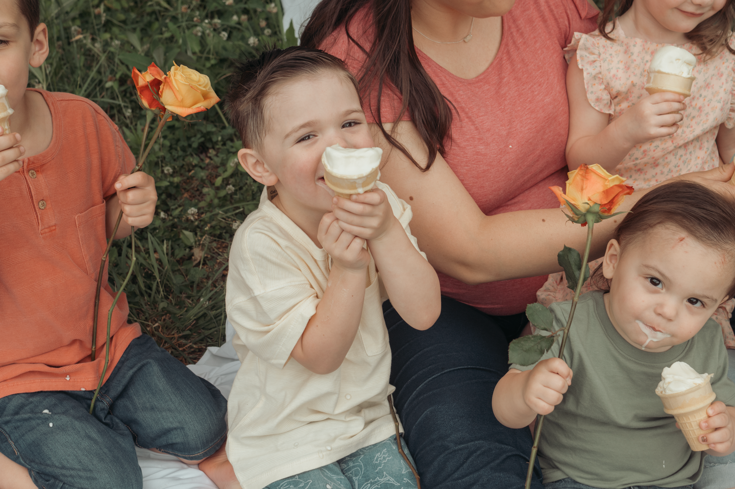 A group enjoys ice cream cones and orange roses while sitting together on grass in warm sunlight.