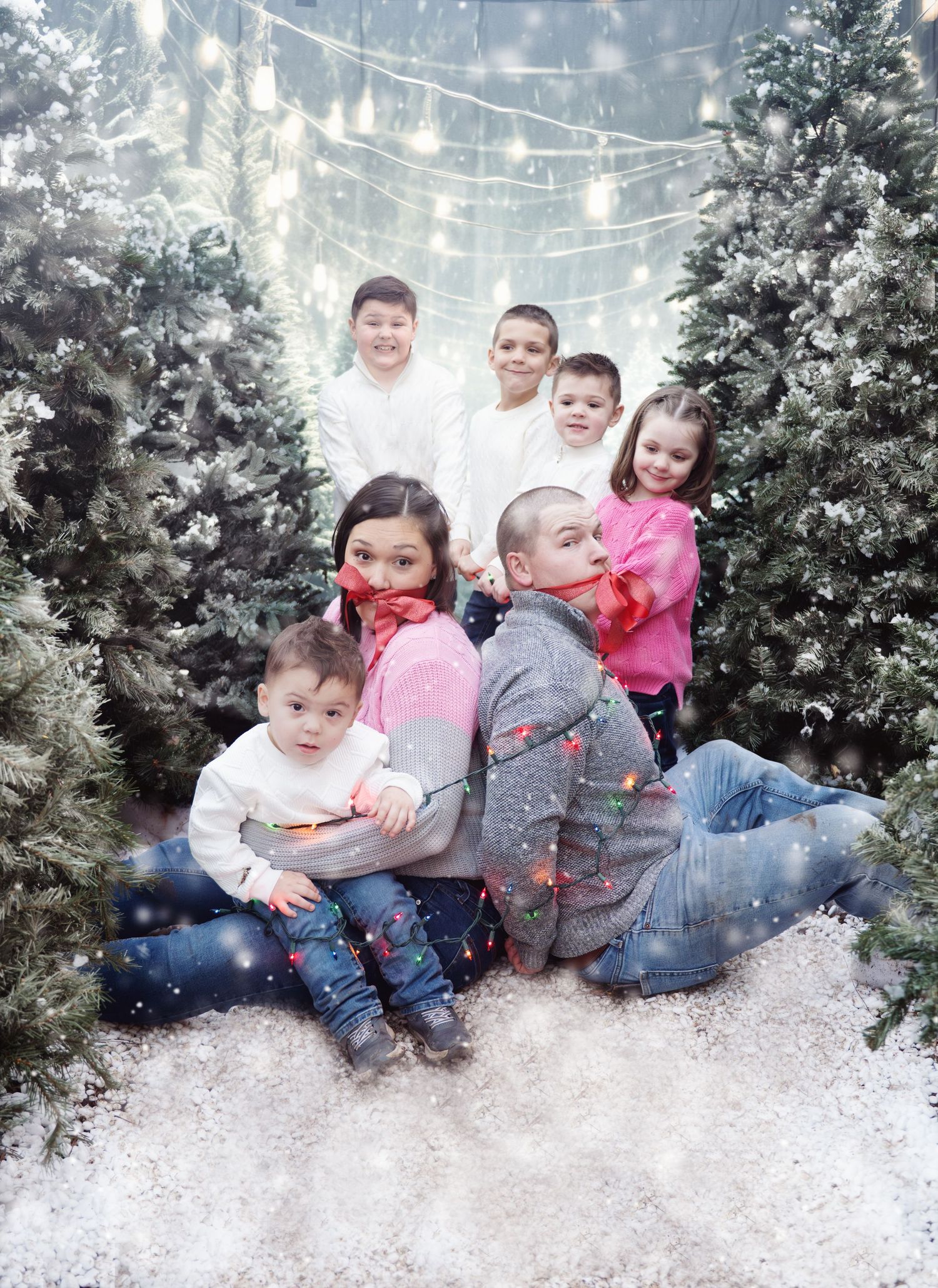 A winter family portrait session with children sitting in artificial snow among decorated evergreen trees and string lights.