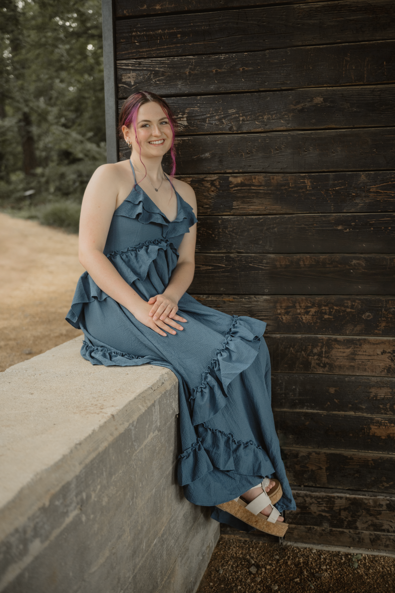Haylee in a flowing blue ruffled maxi dress sits on a concrete ledge against a rustic wooden wall outdoors for her senior session.