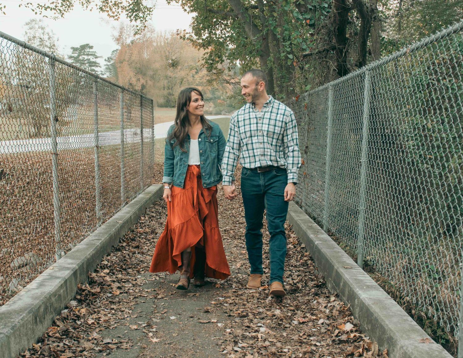 A couple walks hand in hand along a leaf-covered walkway bordered by chain link fences during autumn.