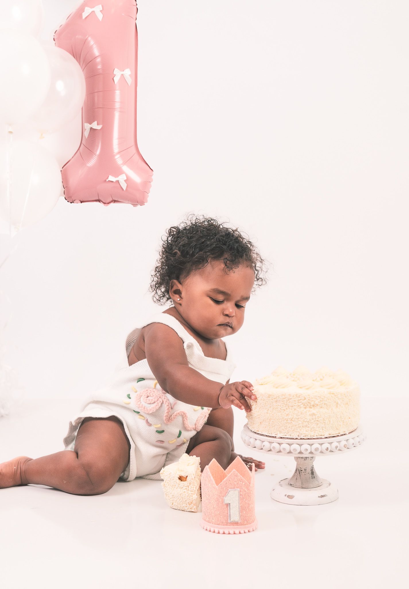 First birthday celebration setup with pink balloon number one, white cake on stand, and crown decoration against light background.