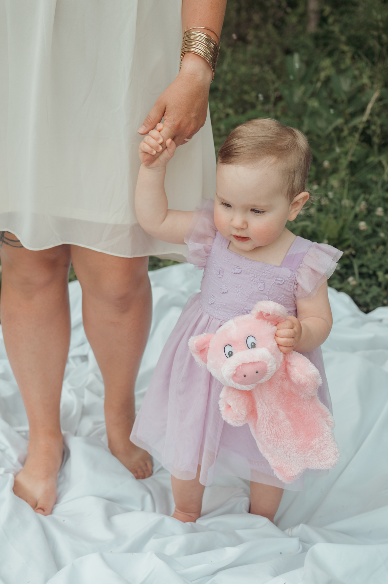A child in a purple dress holds a pink plush toy while standing on white fabric outdoors.