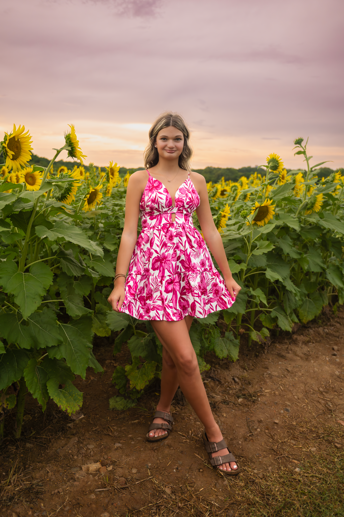 Woman in pink floral dress poses playfully in a sunflower field at sunset against a moody sky.