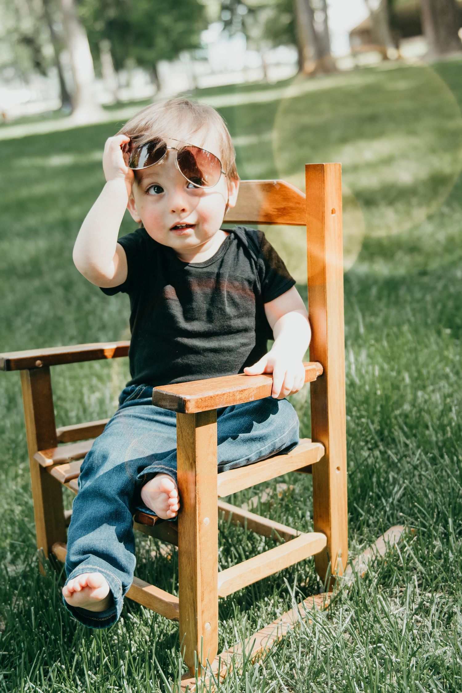Small child wearing sunglasses and black shirt sits in wooden chair on sunny grass lawn.