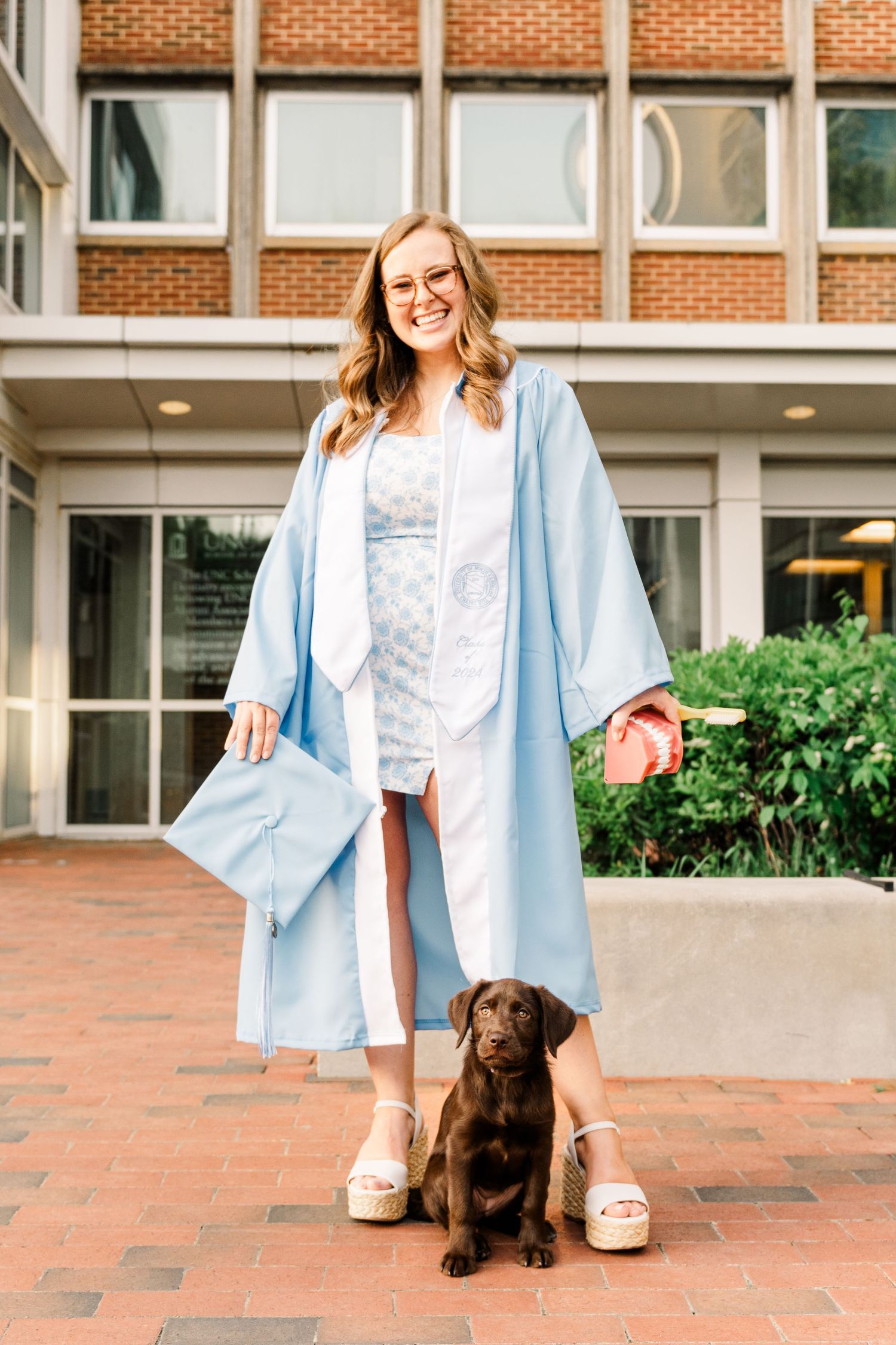 Graduate in light blue cap and gown stands with chocolate lab puppy on brick courtyard of university building.