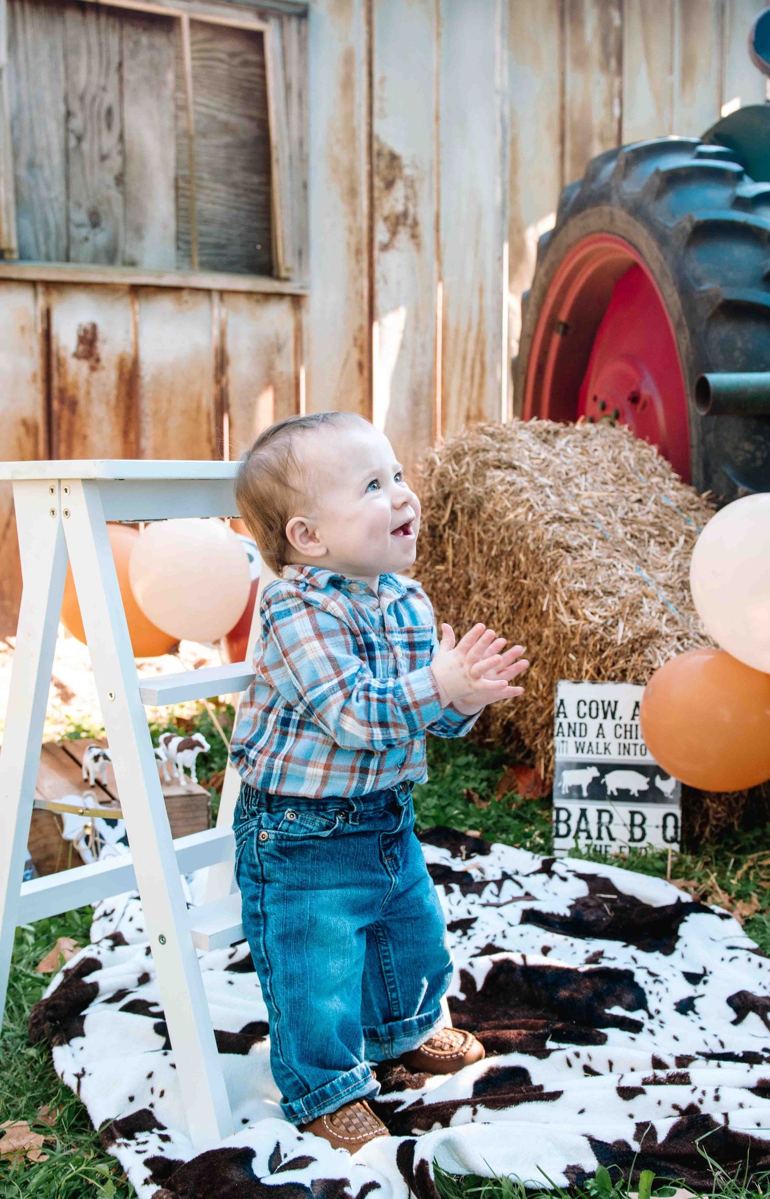 A toddler in plaid shirt and jeans stands excitedly near hay bales and farm decor at a rustic outdoor setting.