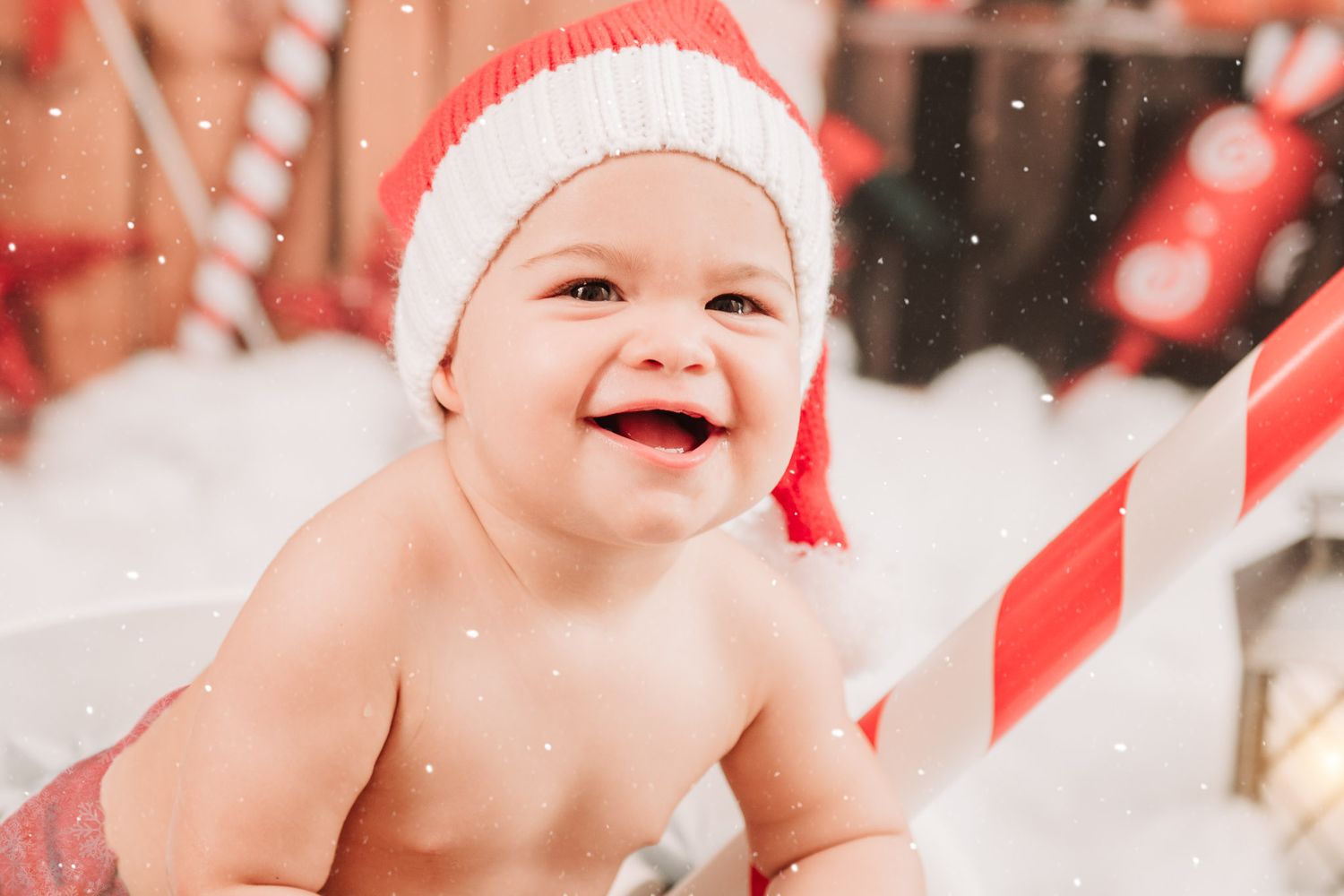 A smiling baby wearing a red and white Santa hat sits amid festive holiday decorations and white snow.