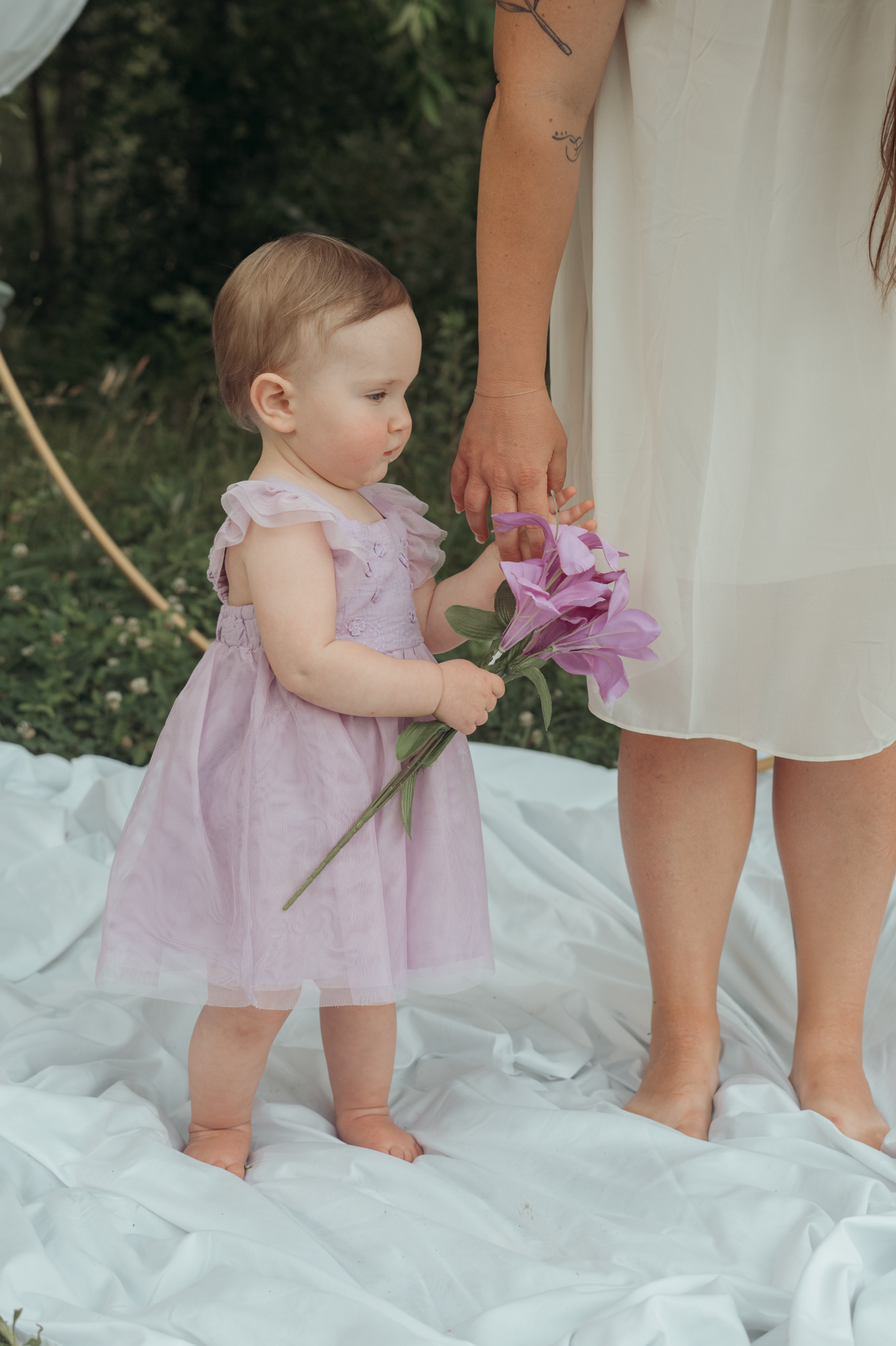 Small child in lavender dress stands on white blanket holding pink flowers beside adult in white dress.