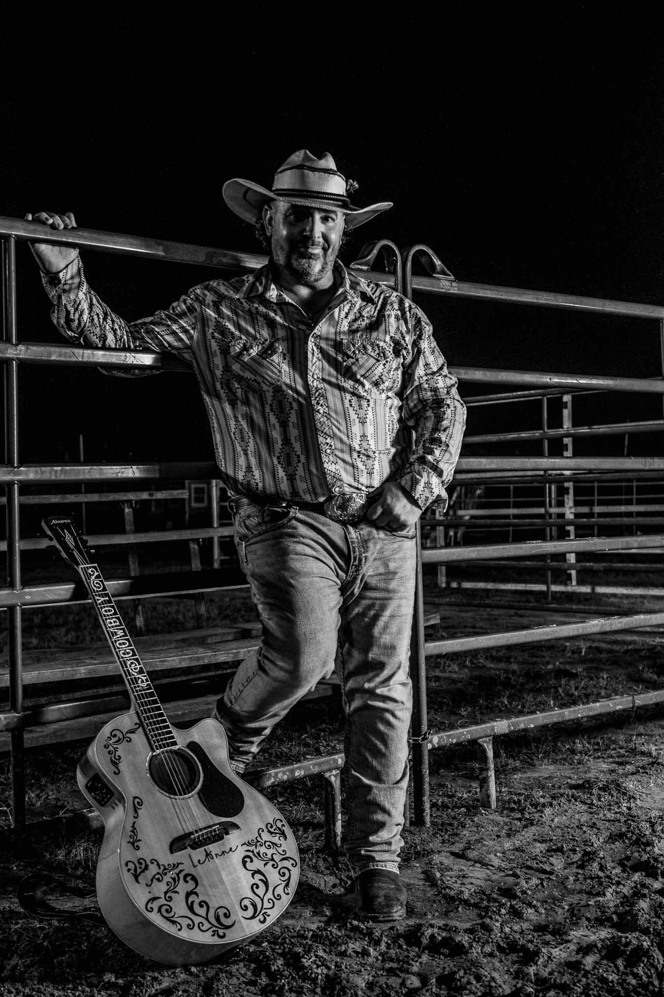 Black and white portrait of a country musician in western attire leaning against fence rails with an acoustic guitar.