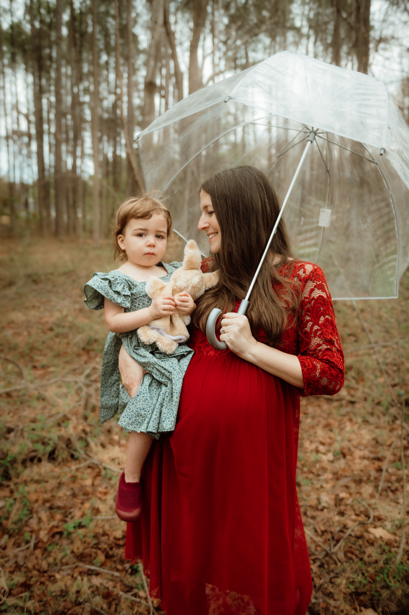 Pregnant woman in red lace dress holds toddler with stuffed animal under clear umbrella in forest.