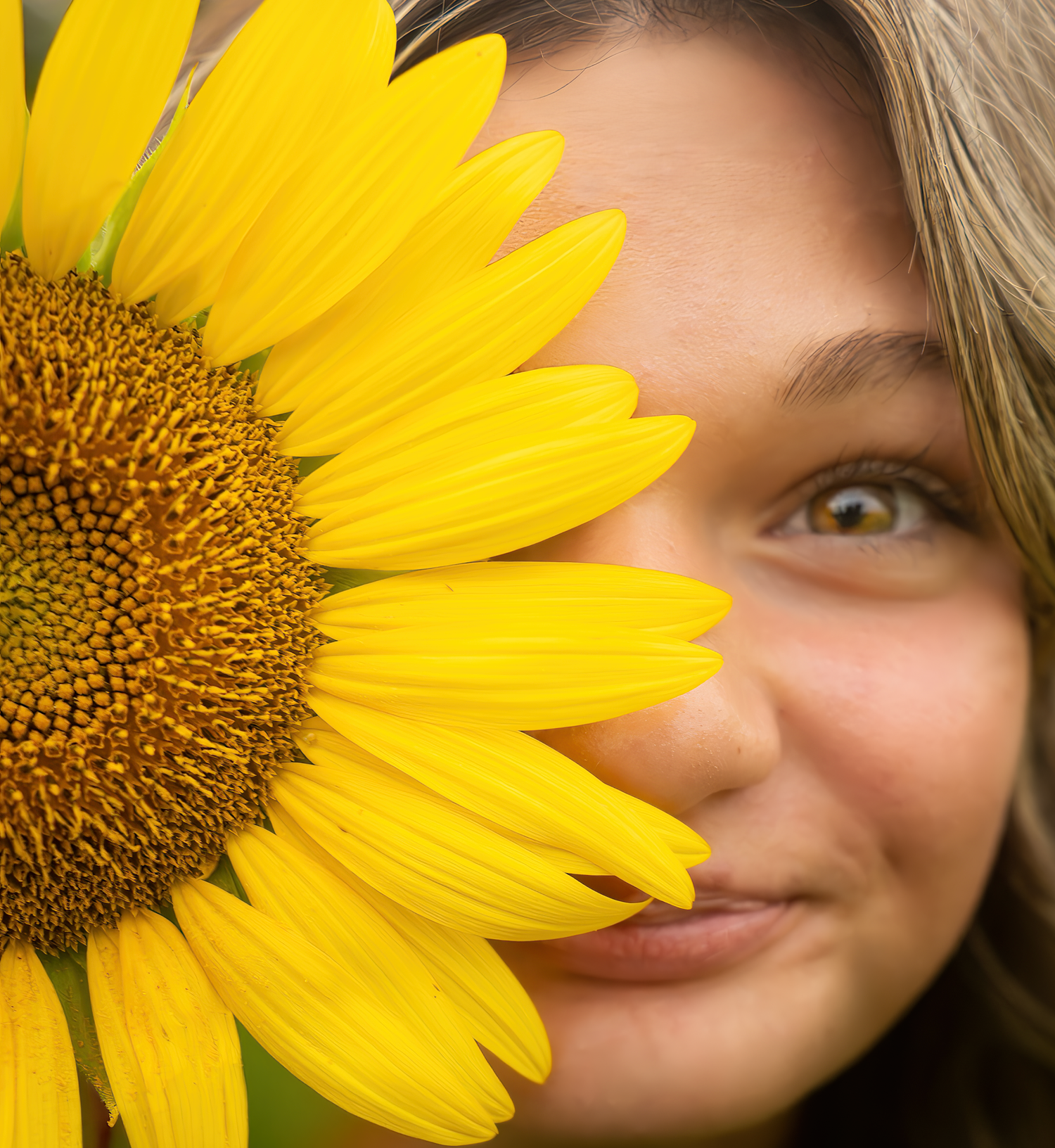 A bright yellow sunflower petal frames an eye and partial face peeking out from behind the large bloom.