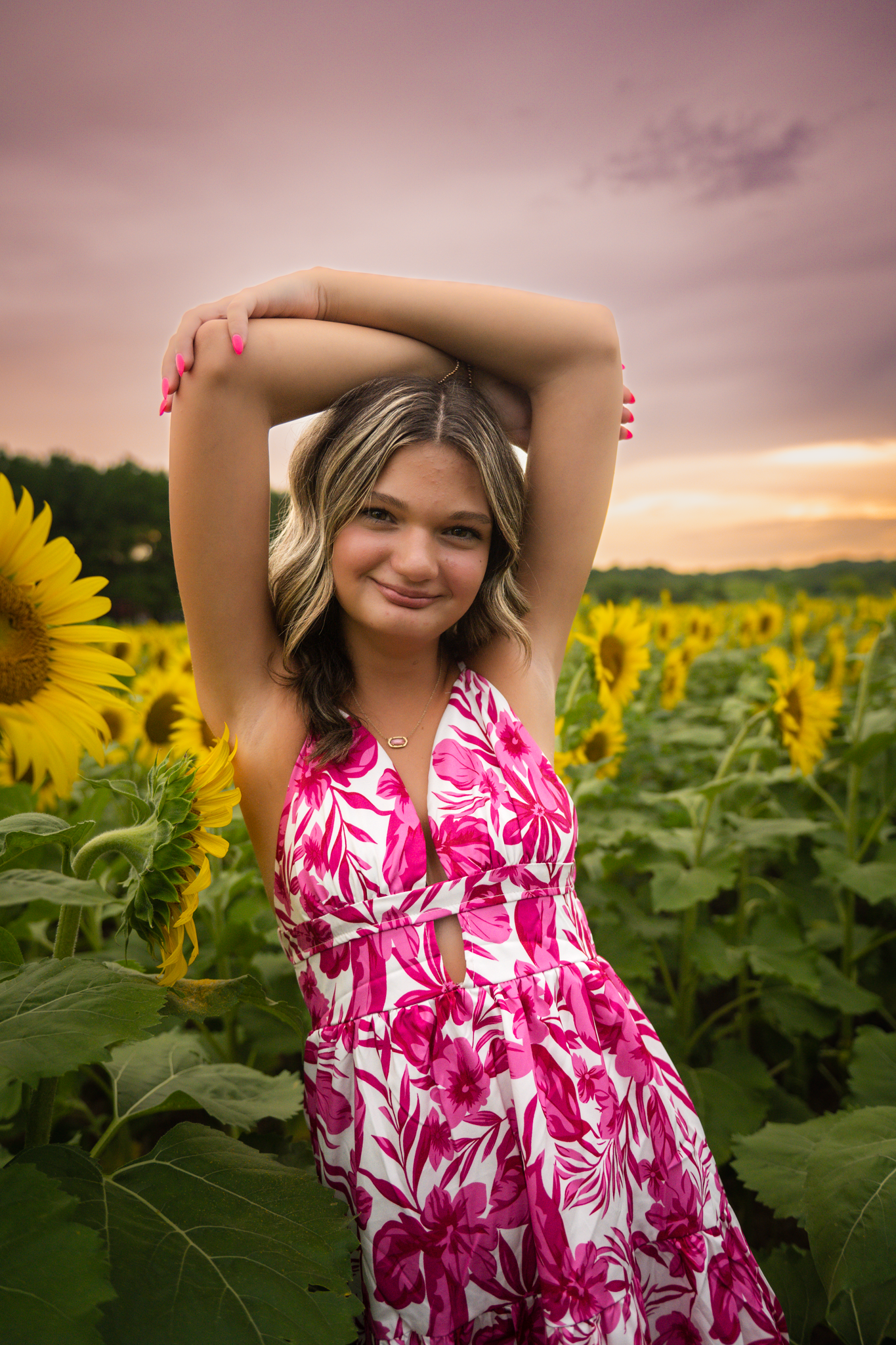 A person in a pink floral dress poses in a sunflower field at sunset with arms raised overhead.
