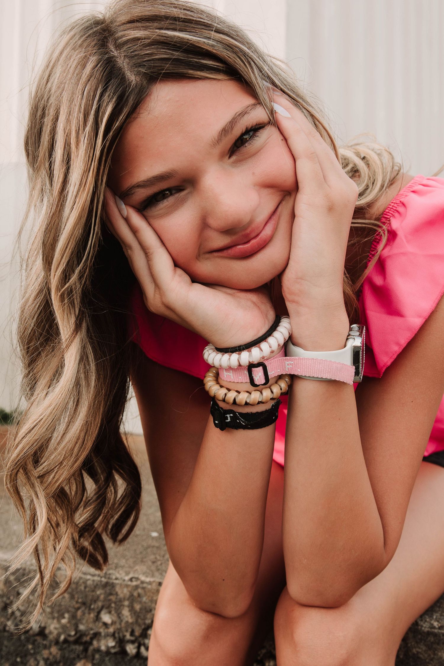 Smiling person in pink top wearing stacked beaded bracelets rests head in hands while posing outdoors.