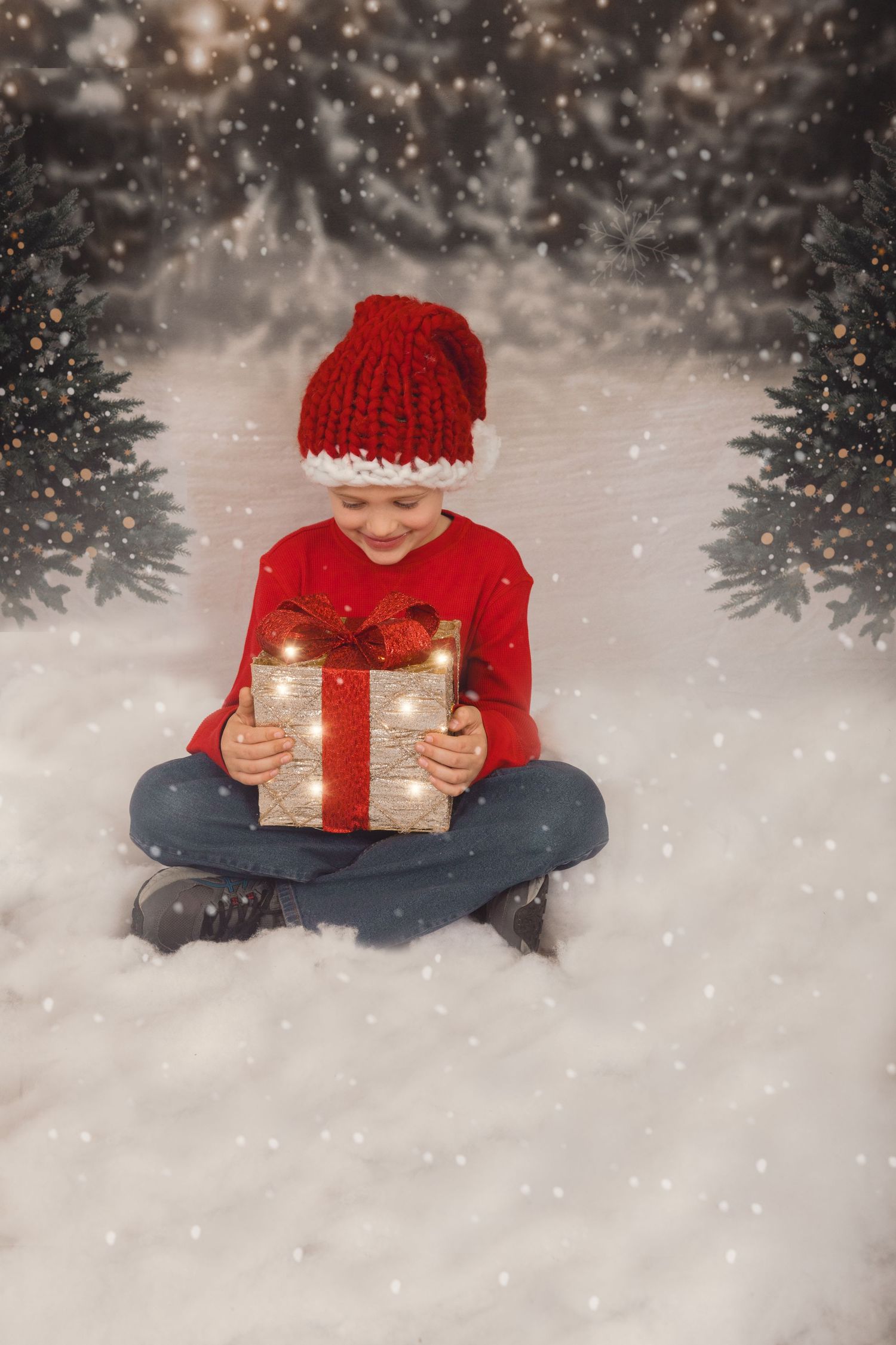 A child in a red knit hat and sweater sits in snow holding an illuminated Christmas present while snowflakes fall.