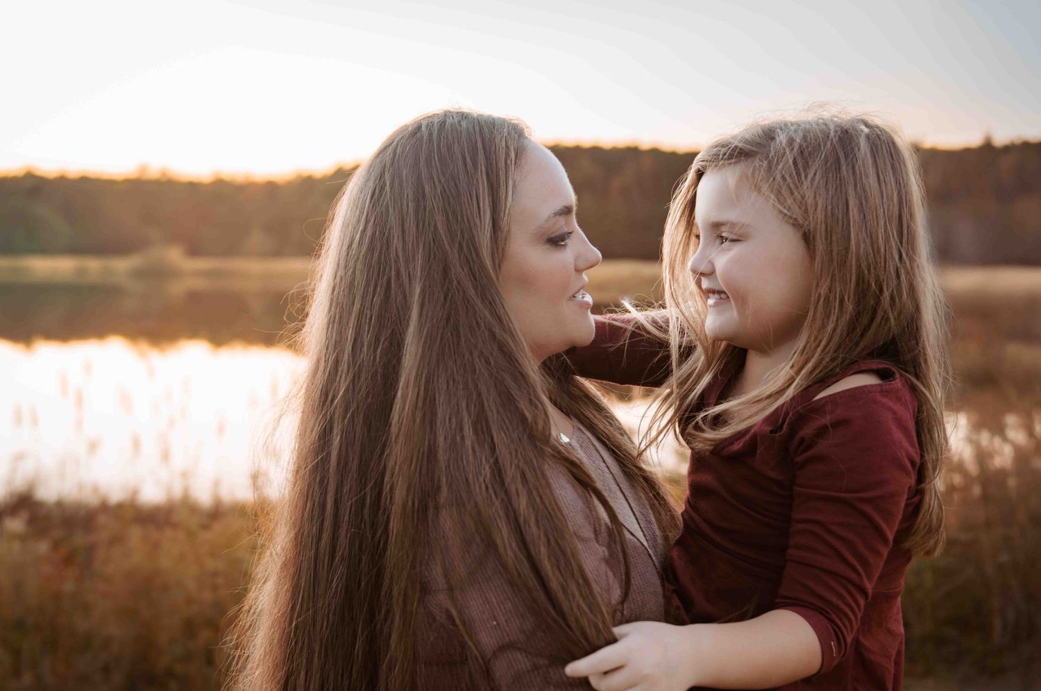 A parent and child share a tender moment at sunset by a lake while wearing matching burgundy tops.