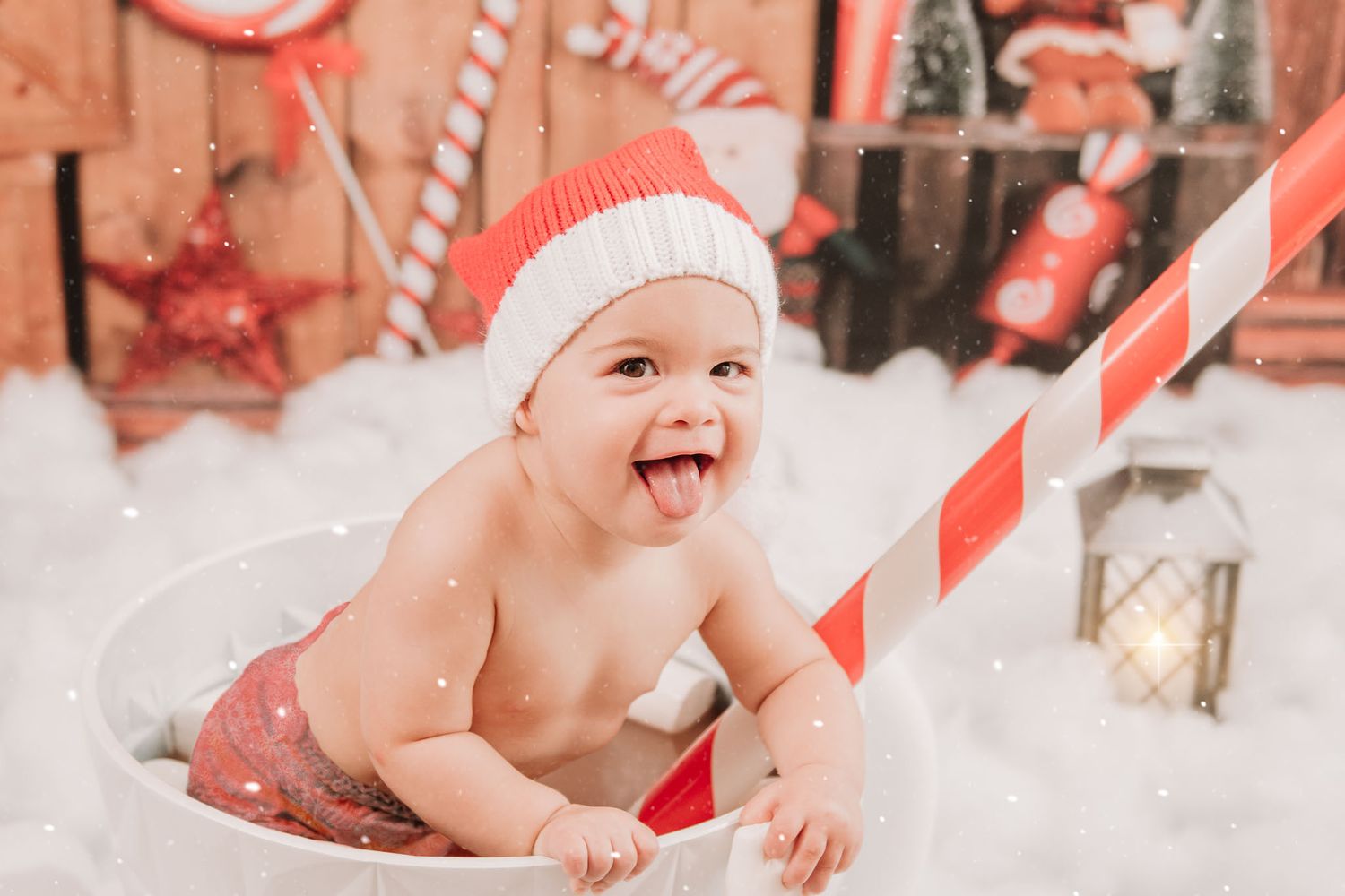Baby in Santa hat lying on white fluffy blanket with candy cane striped prop during Christmas photo session.