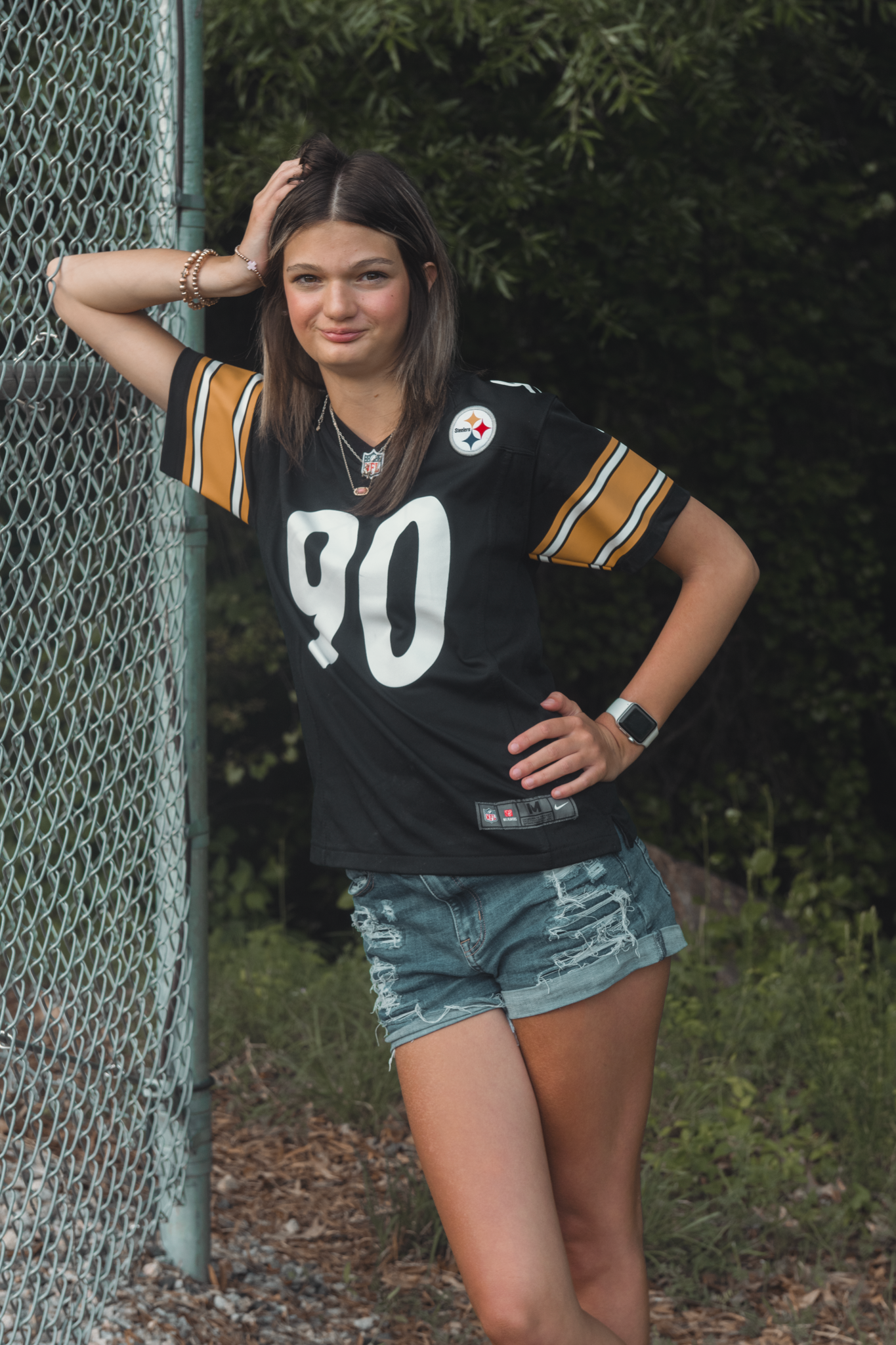 A person in a Pittsburgh Steelers jersey number 90 poses by a chain link fence while wearing denim shorts.