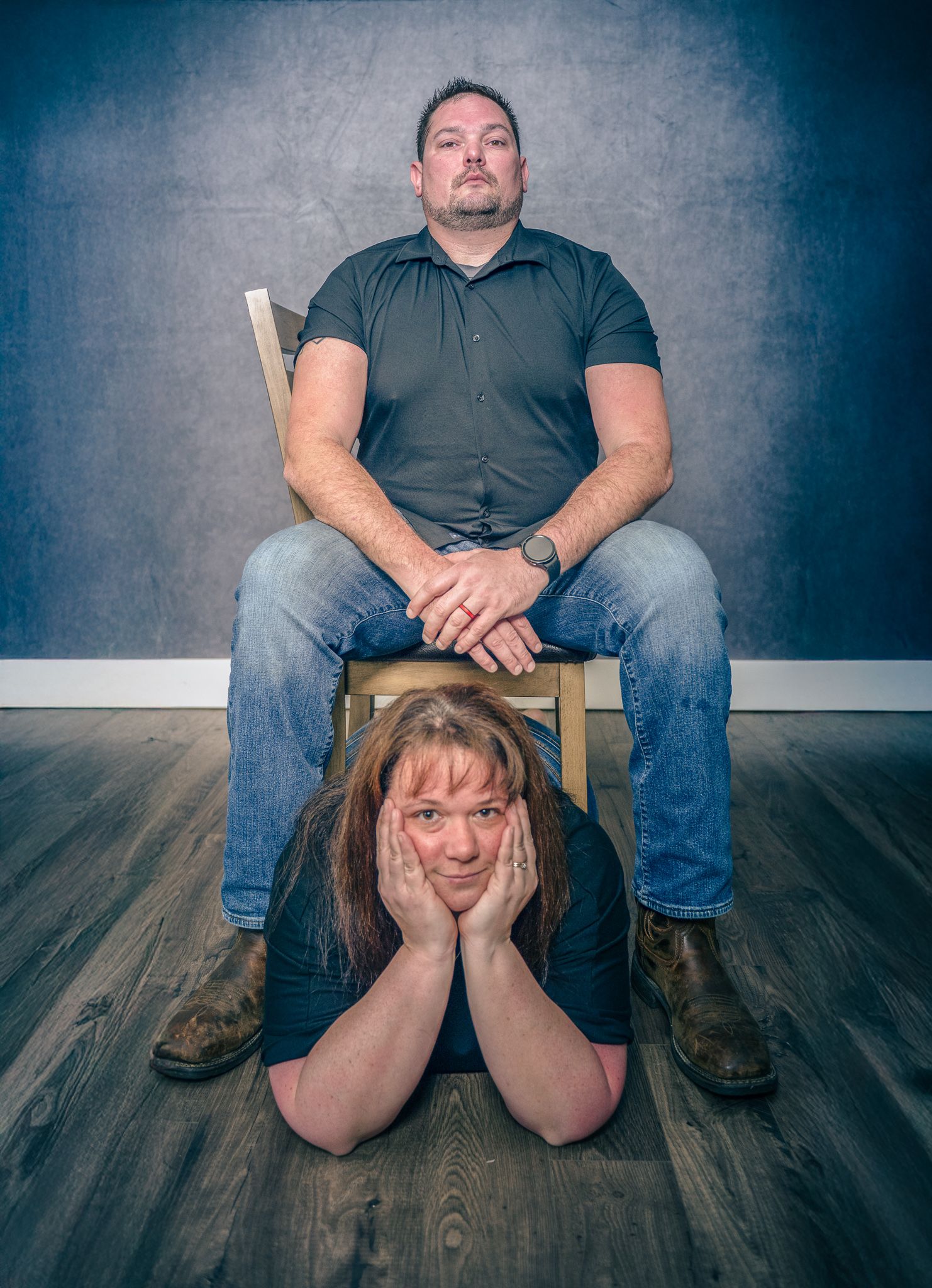 A playful portrait of two people with one sitting on a wooden chair while another poses beneath on the floor.