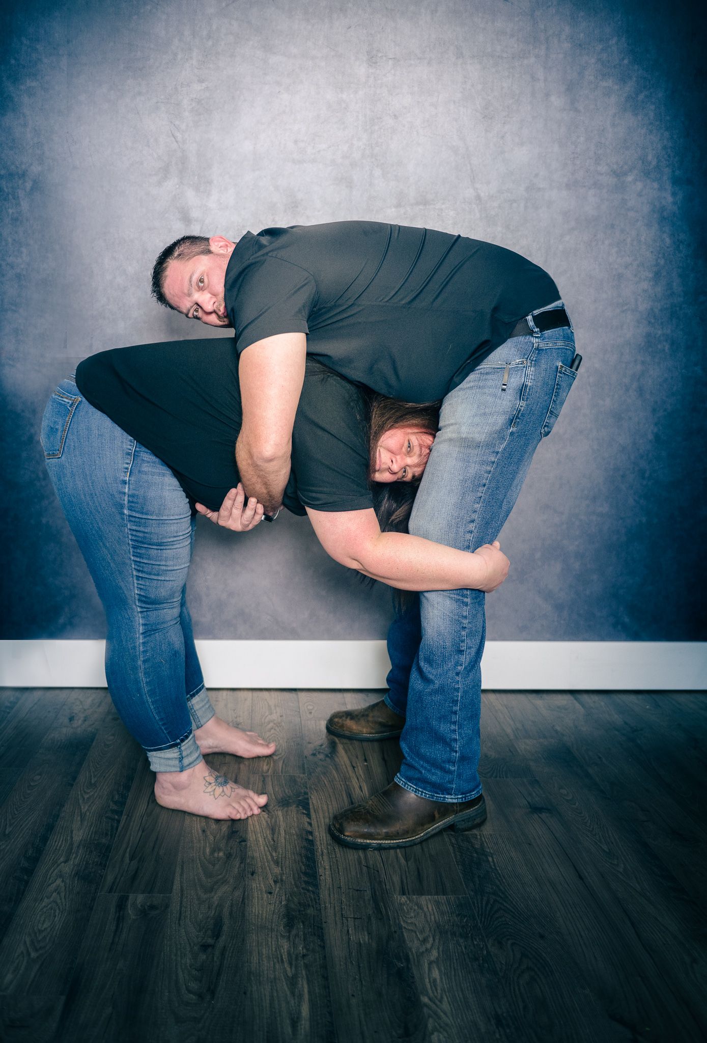 Two people in jeans demonstrating a wrestling move against a gray backdrop on dark hardwood flooring.