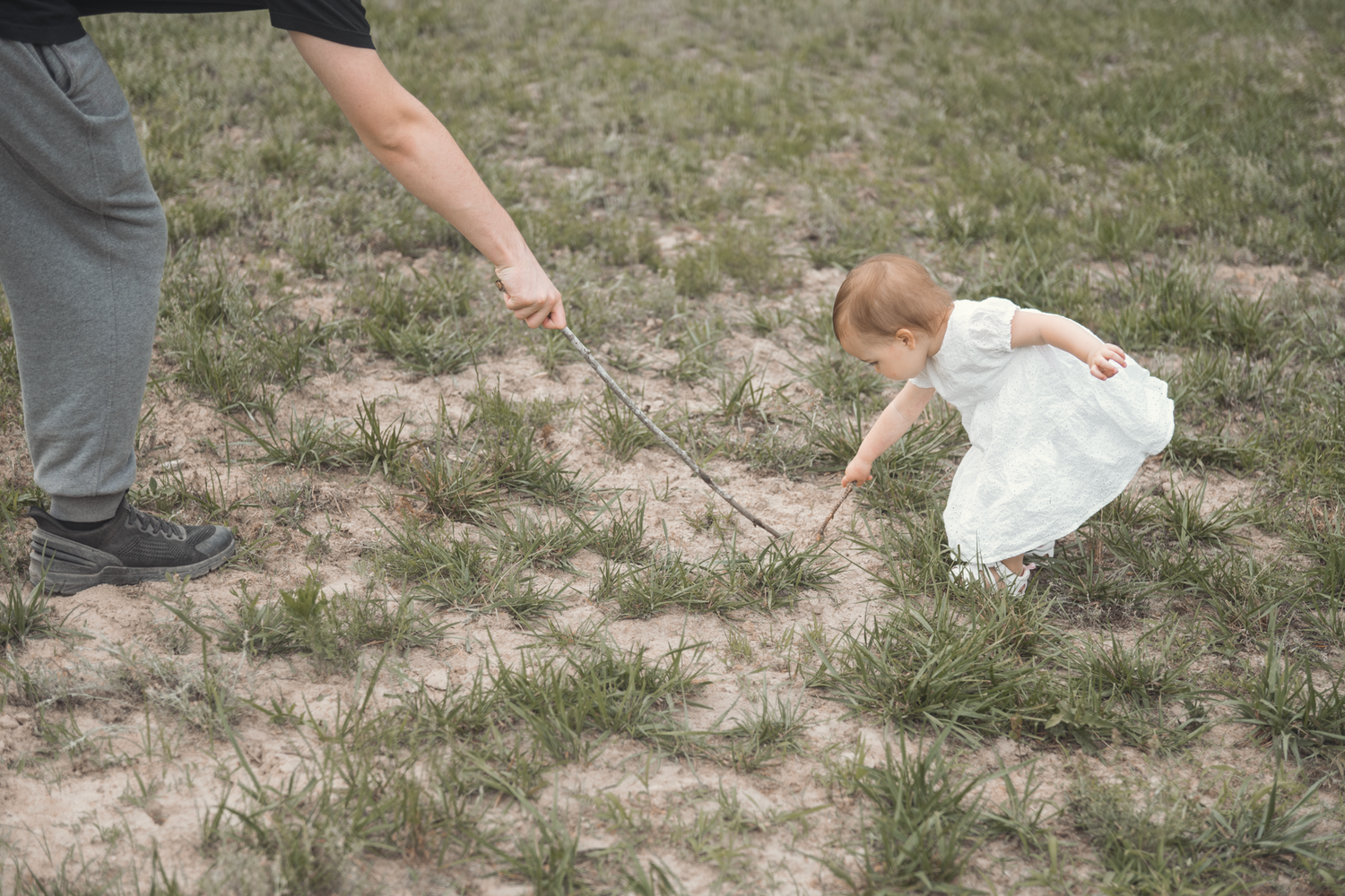 A small child in a white dress bends down to touch the dry ground while an adult's hand points nearby.