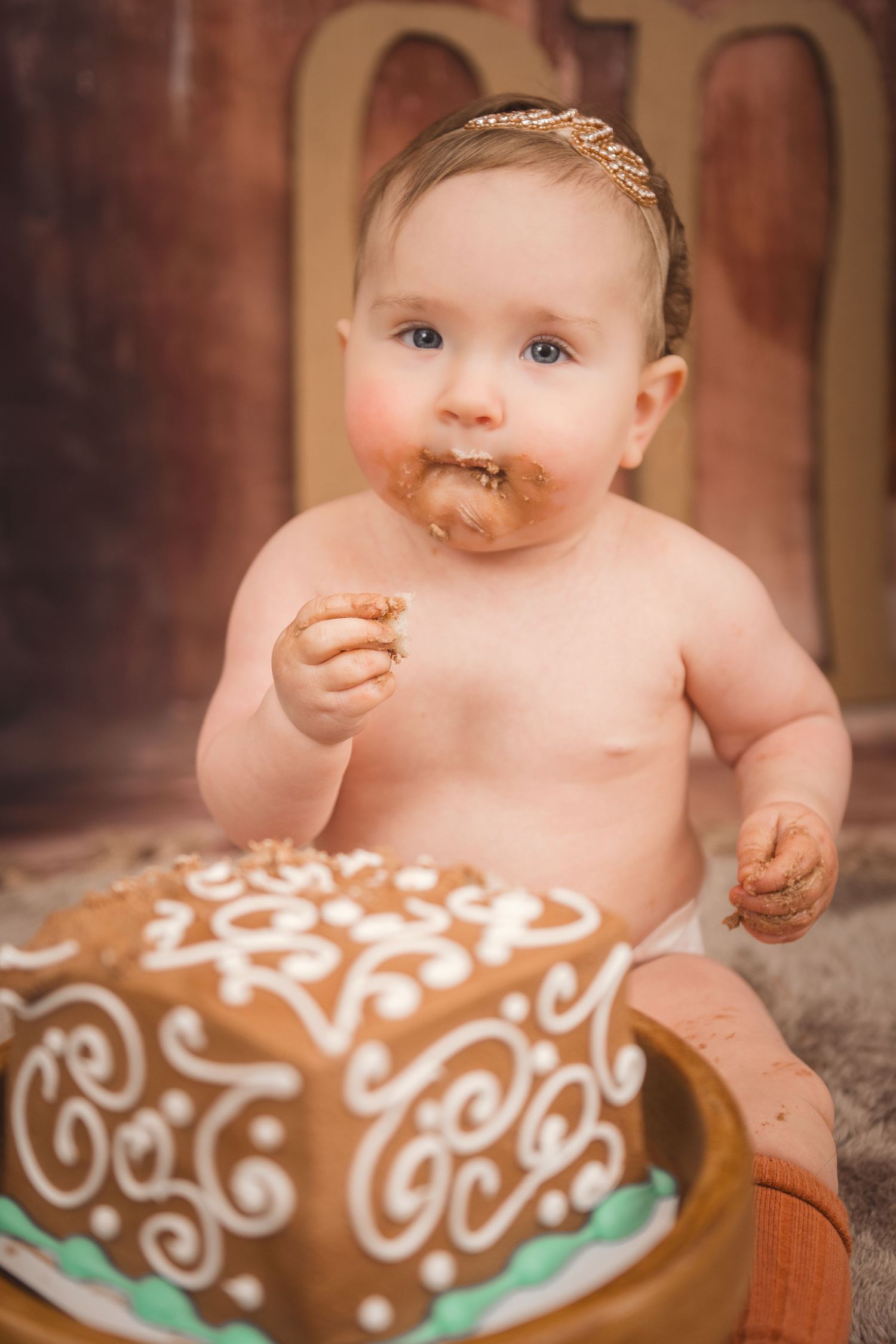 A decorated brown cake with white frosted swirls sits in front of a messy baby enjoying a sweet treat.