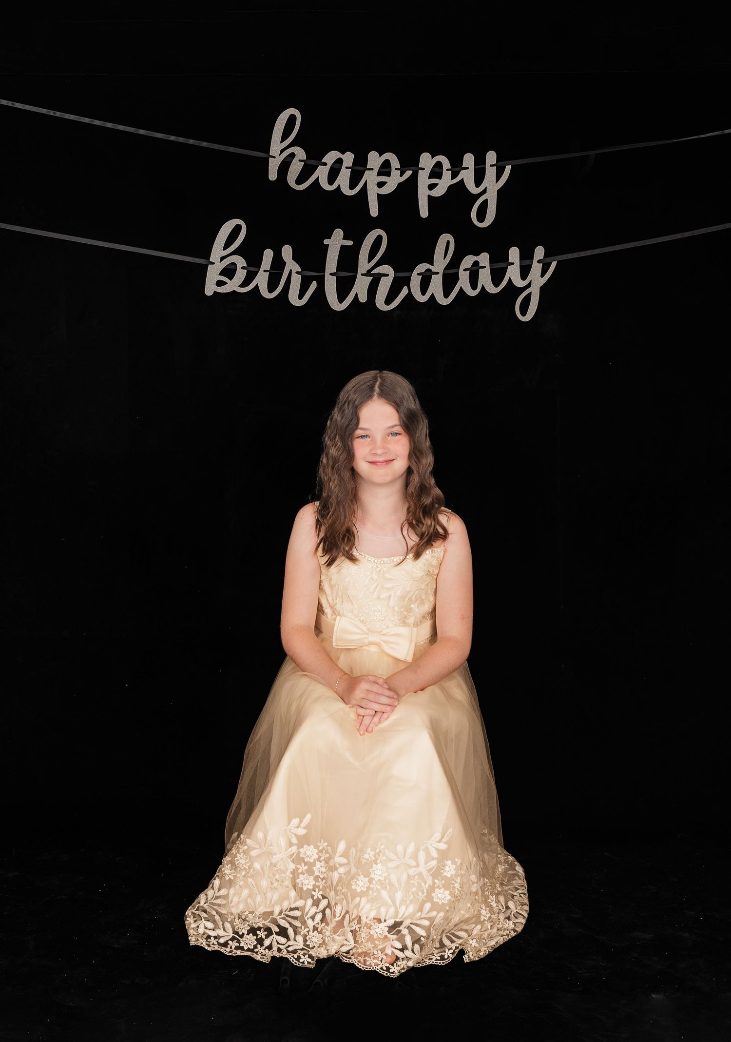 A formal celebration photo with 'happy birthday' text hanging above featuring a cream-colored dress against a black background.