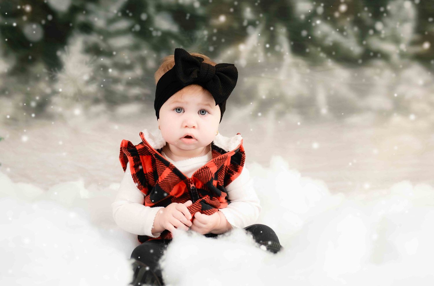 A baby in buffalo plaid vest and black bow sits in artificial snow with evergreens in background.